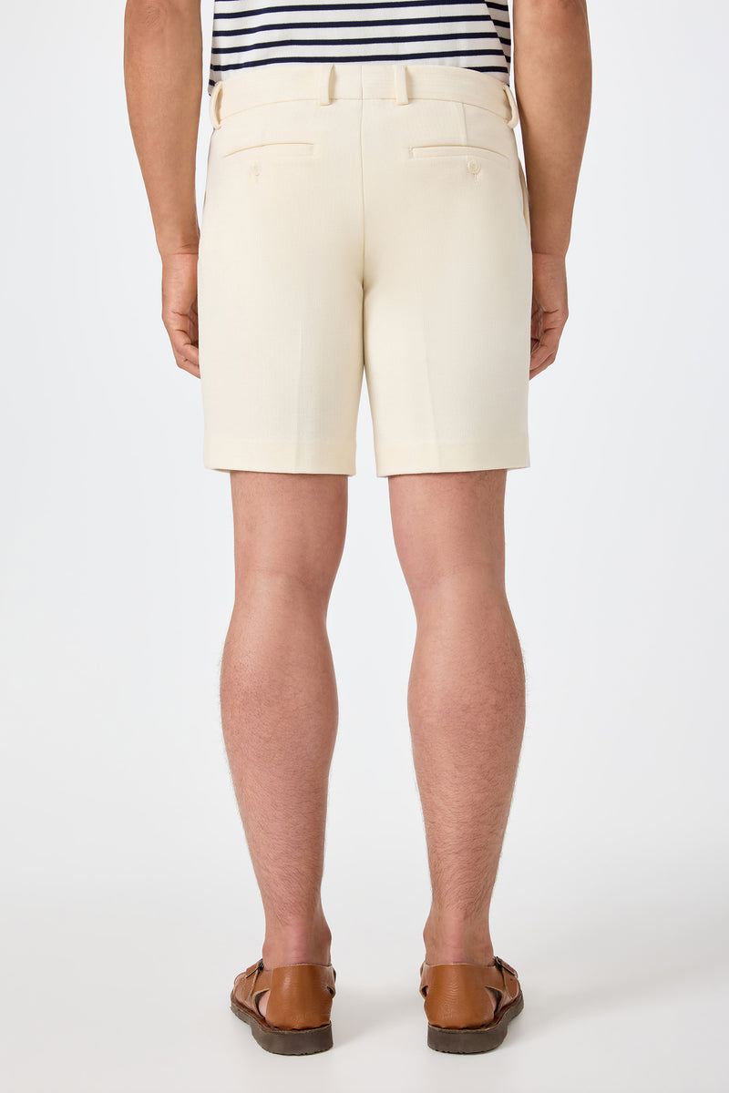 A person stands facing away in the Lawrence Short - Ecru, paired with a striped tucked-in shirt and brown loafers against a plain white background.