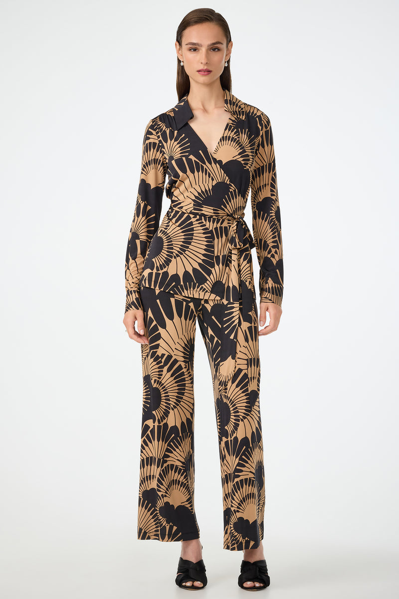 A woman faces forward, wearing the Makeba Wrap Top in Juniper Jersey— a long-sleeve beige and black wrap top with an abstract leaf pattern—paired with matching wide-leg pants. She has straight brown hair and black open-toe heels.
