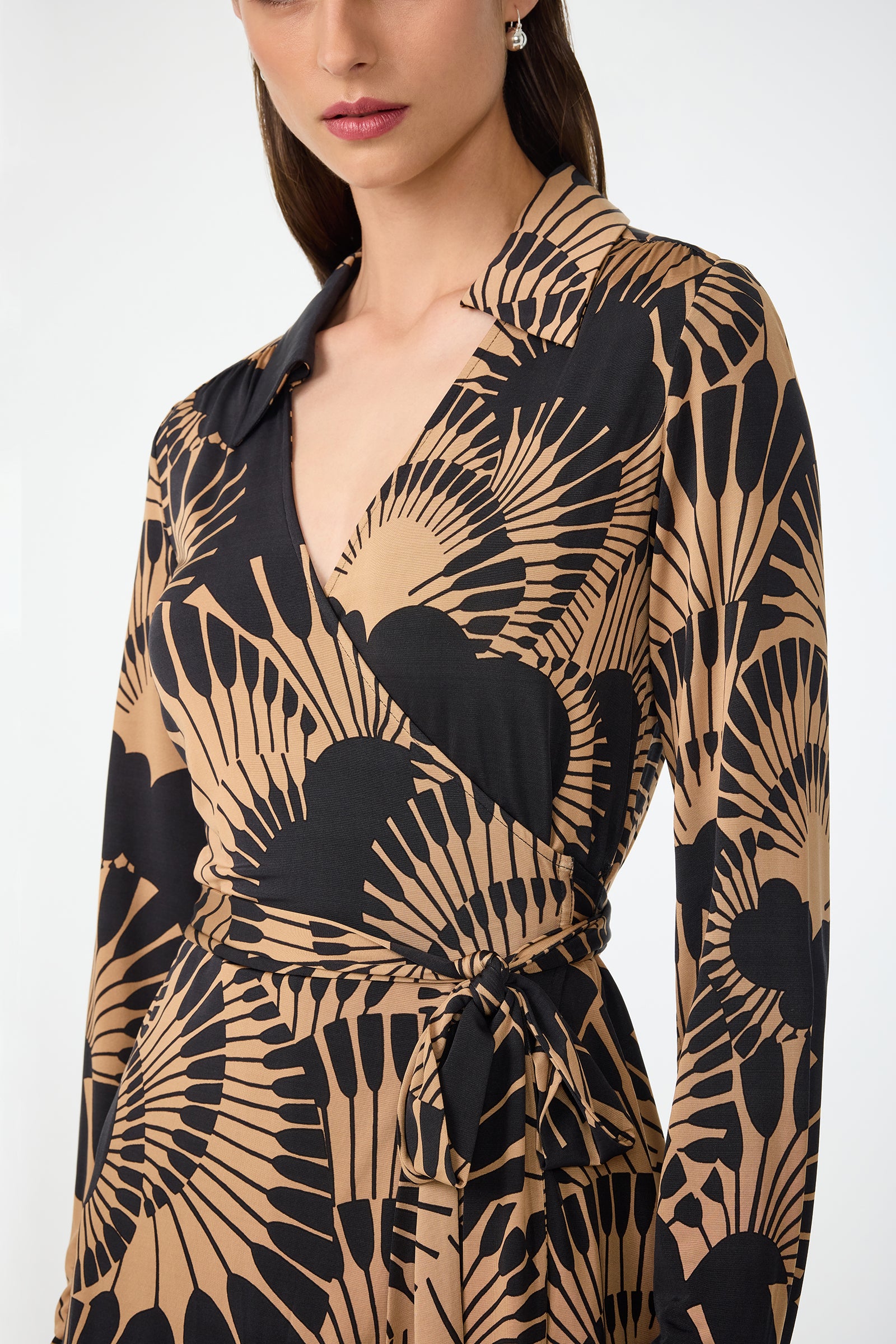 A woman wearing the Makeba Wrap Top in Juniper Jersey, featuring long sleeves, a black and tan fan pattern, V-neckline, tied waist, and collar. The image shows her from shoulders to mid-thigh.