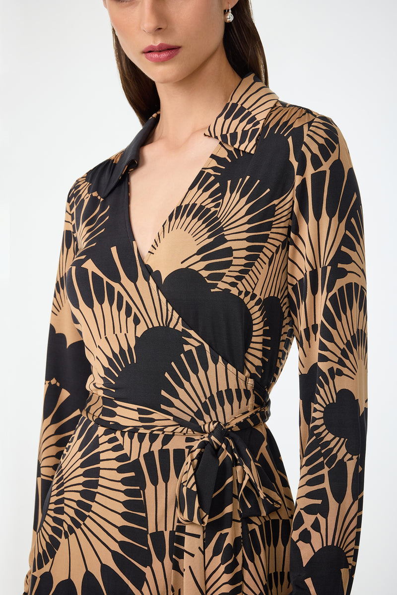 A woman wearing the Makeba Wrap Top in Juniper Jersey, featuring long sleeves, a black and tan fan pattern, V-neckline, tied waist, and collar. The image shows her from shoulders to mid-thigh.