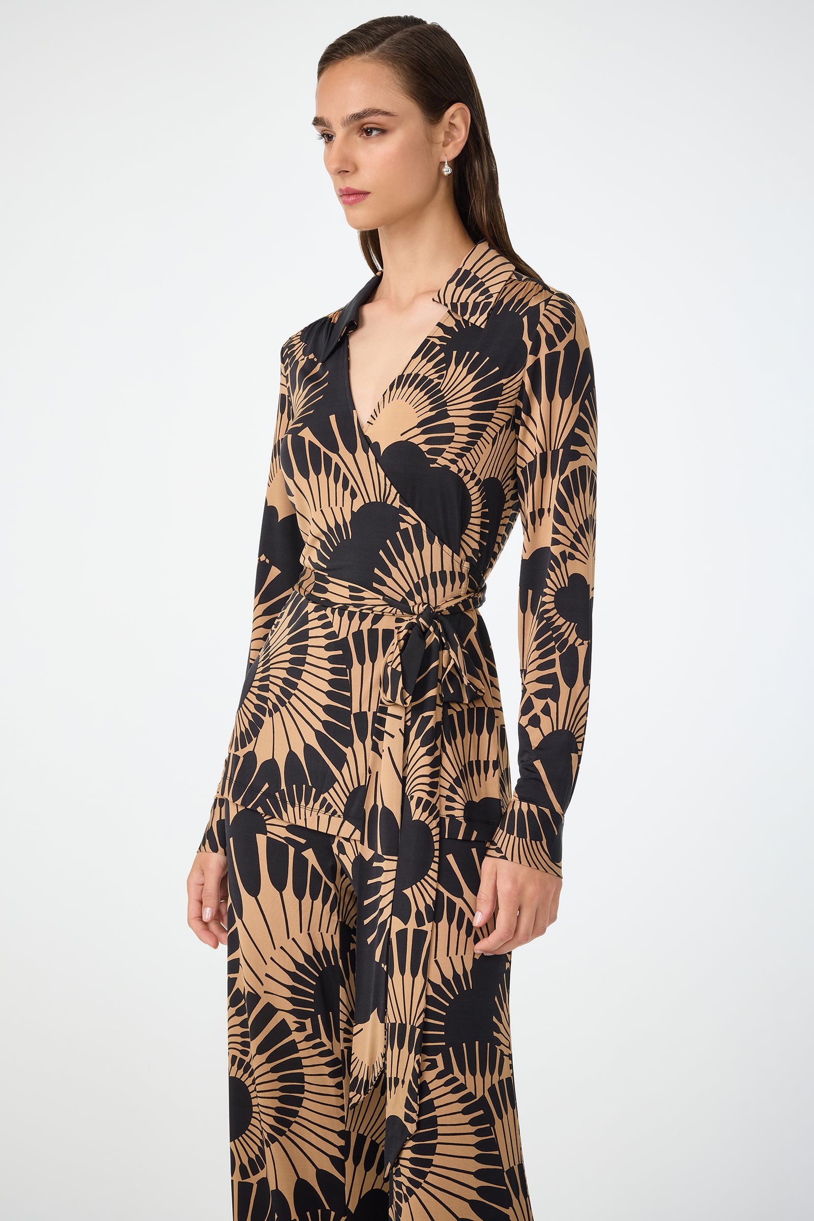 A woman wears the Makeba Wrap Top in Juniper Jersey, featuring long sleeves, a beige and black pattern, and a tie at the waist, standing against a plain white background.