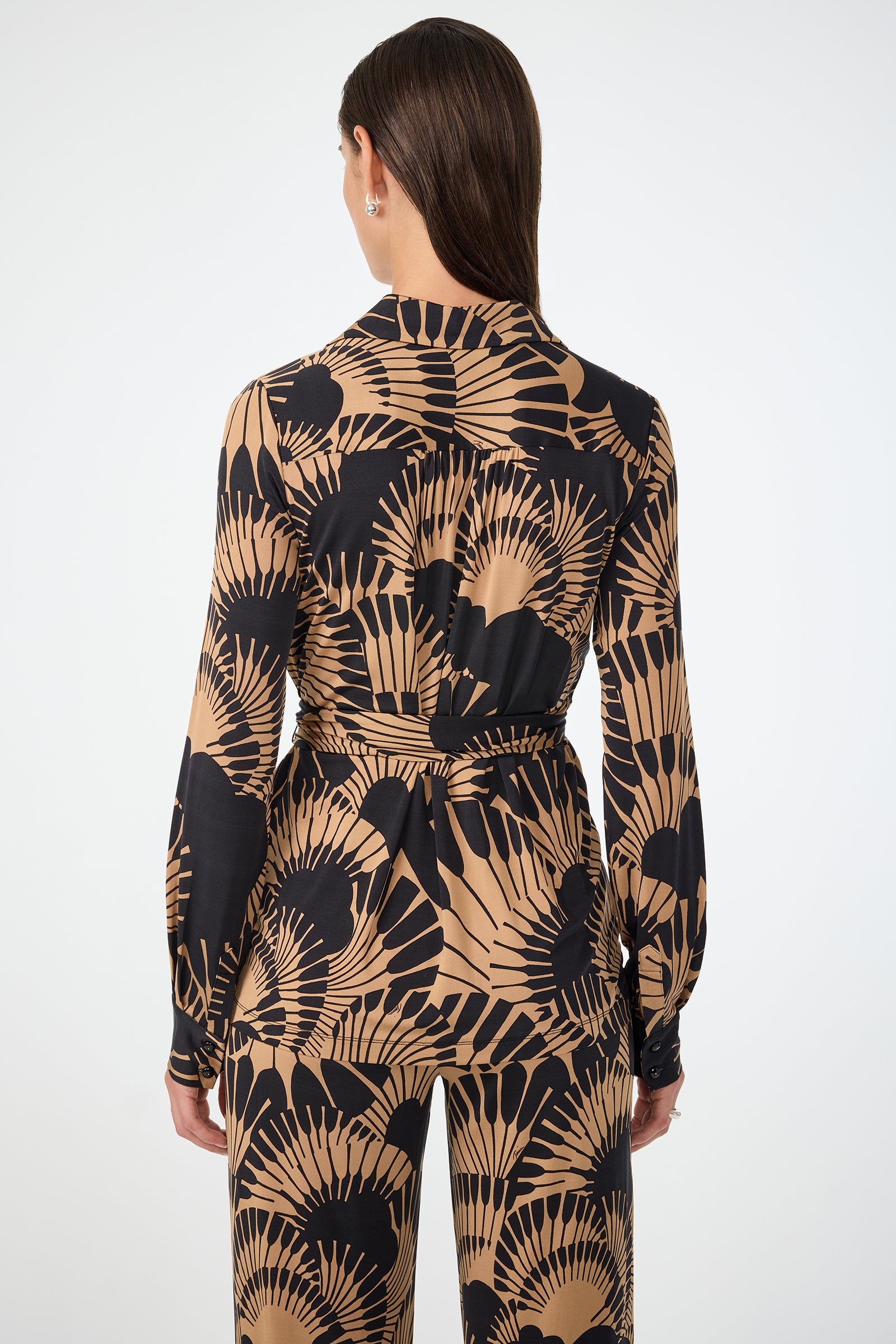 A woman with straight brown hair stands facing away, wearing the Makeba Wrap Top in Juniper Jersey. The top features a tan and black fan pattern, a waist tie, and is styled with matching pants against a plain white background.