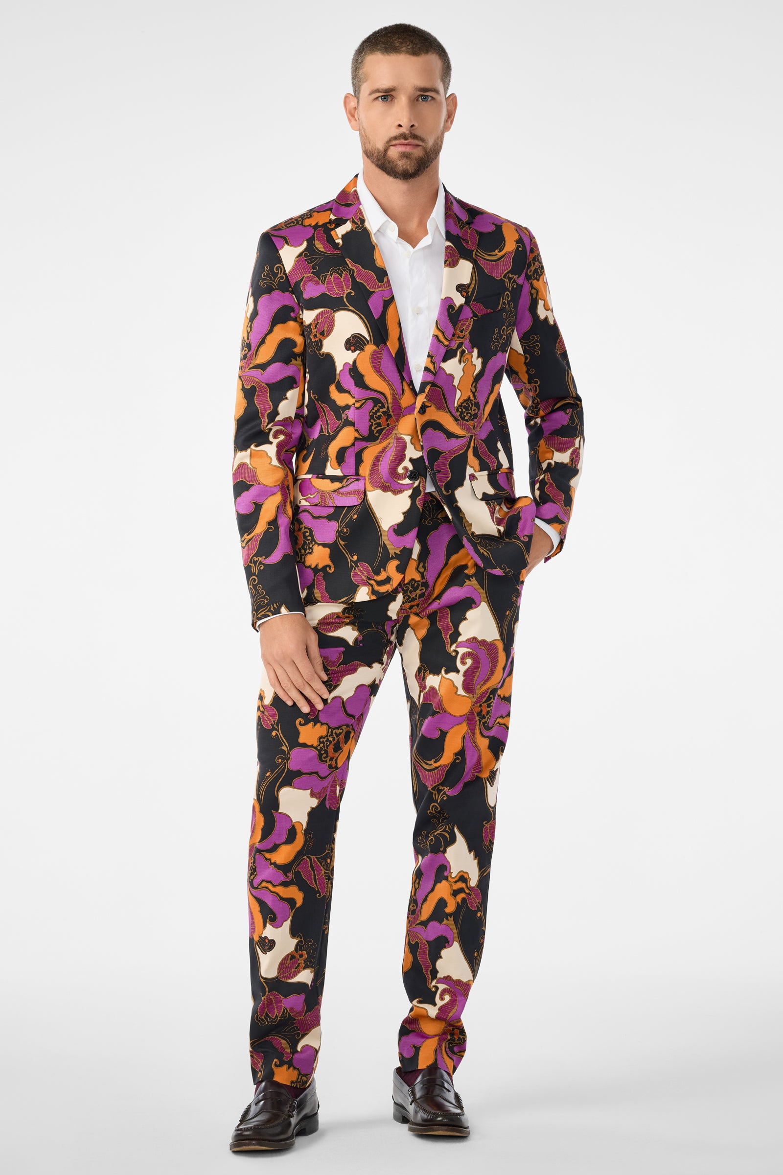 A man with short hair and a trimmed beard faces forward wearing the Thurston Blazer, featuring a bold abstract pattern in black, purple, pink, and orange over a white dress shirt and dark shoes.