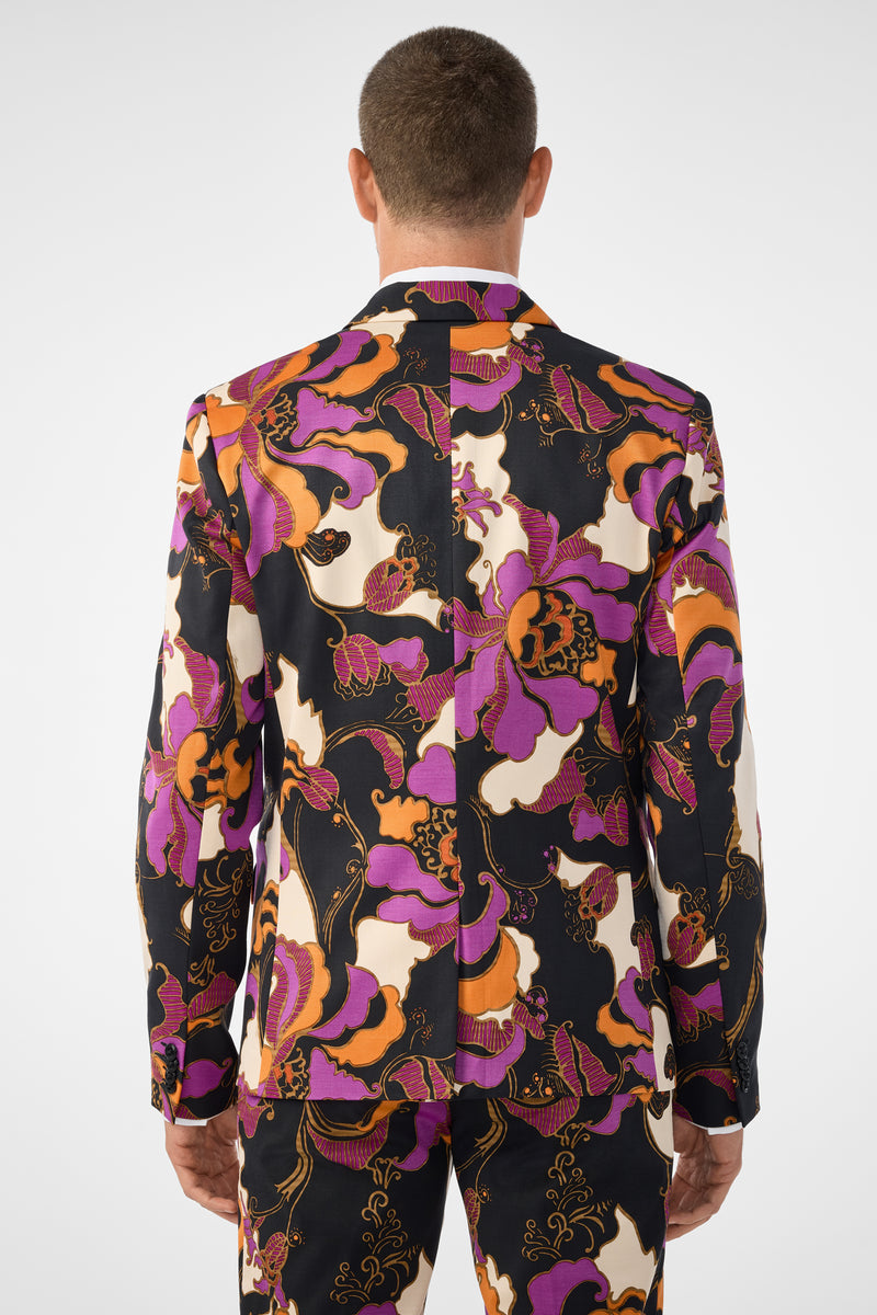 A man is shown from behind wearing the Thurston Blazer, a colorful suit jacket and matching pants with a bold, abstract floral pattern in orange, purple, black, and white. The background is plain white.