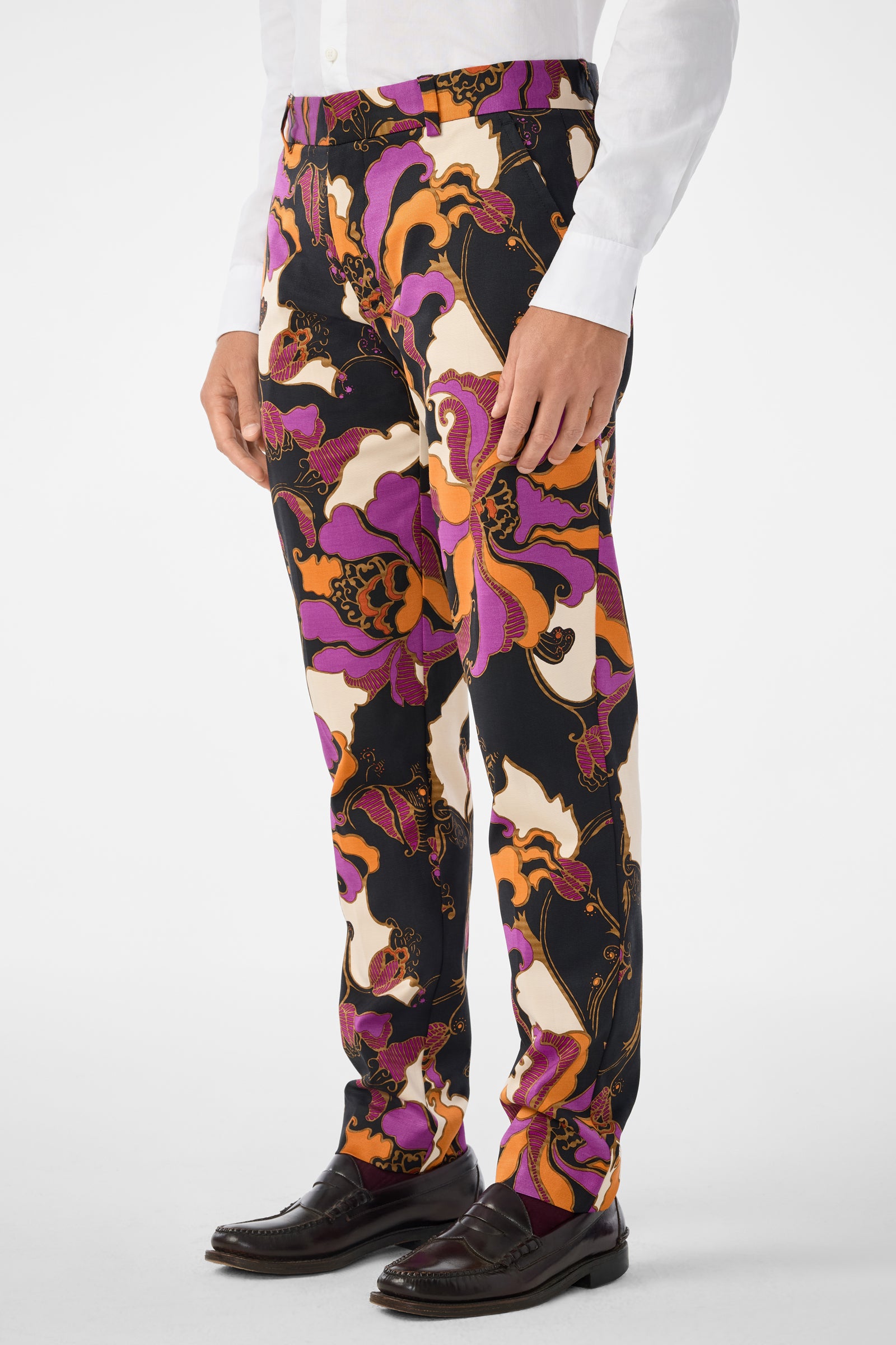 A person wearing Clyde Slim Trouser—colorful, abstract-patterned pants in purple, orange, black, and cream—paired with a white shirt and dark brown dress shoes. The focus is on the trousers.