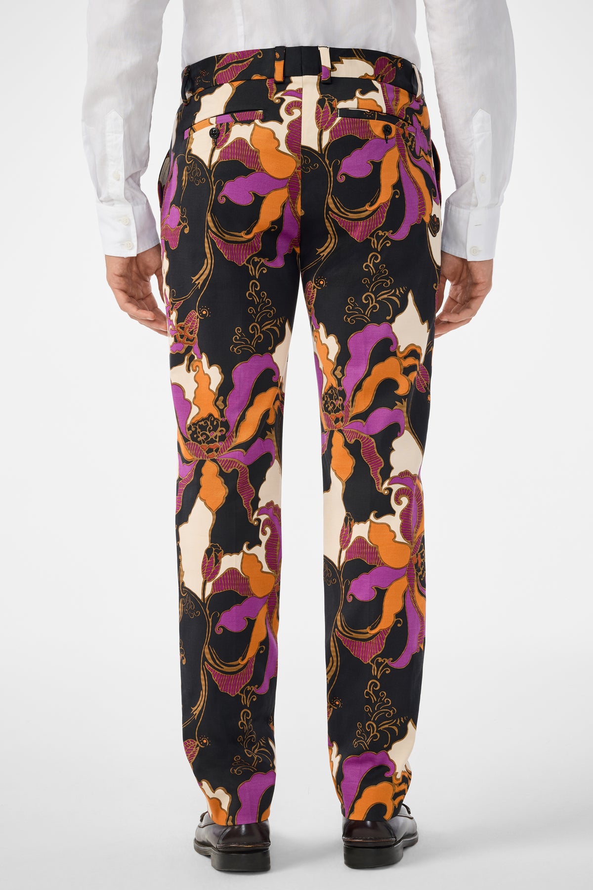 A person is shown from the back against a plain light background, wearing black dress shoes, a white dress shirt, and Clyde Slim Trouser featuring colorful patterns in orange, purple, black, and cream.