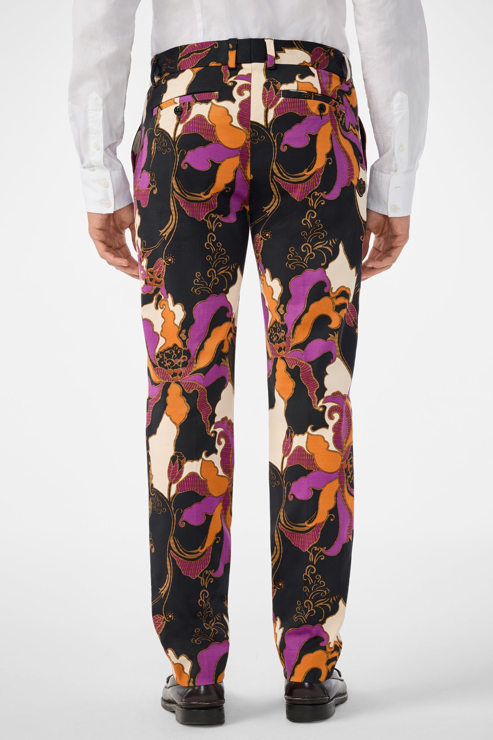 A person is shown from the back against a plain light background, wearing black dress shoes, a white dress shirt, and Clyde Slim Trouser featuring colorful patterns in orange, purple, black, and cream.