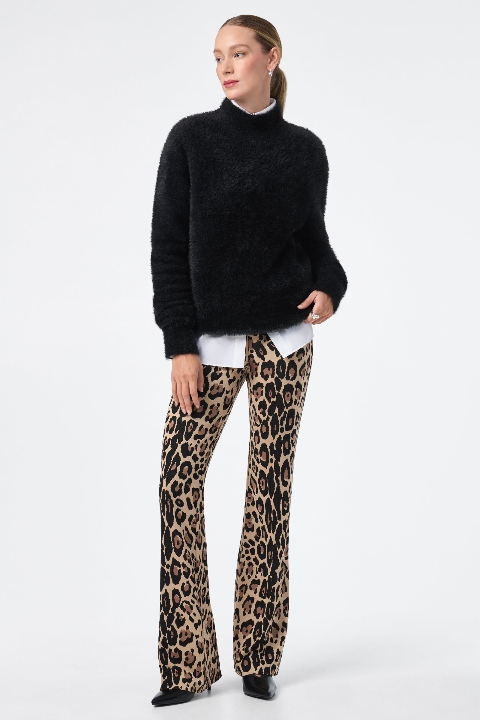 A woman wears the Litsa Button-Up Cotton Shirt under a fuzzy black sweater, paired with flared leopard-print pants and black shoes, standing against a plain white background.