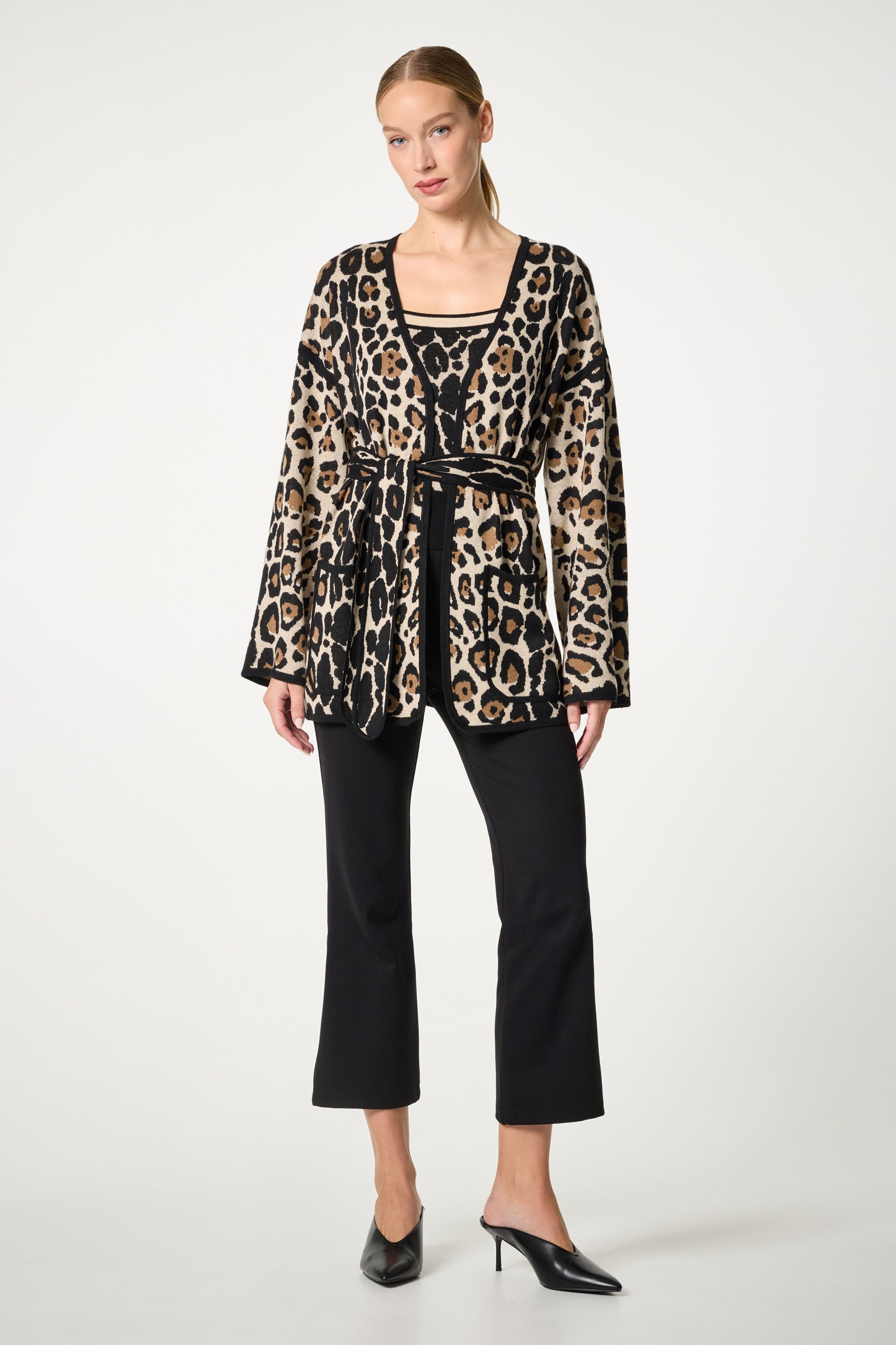 A woman poses in a studio against a plain background, wearing the Wright Wrap Cardigan with a black belt, a matching top, black cropped pants, and black heeled mules. Her hair is pulled back.