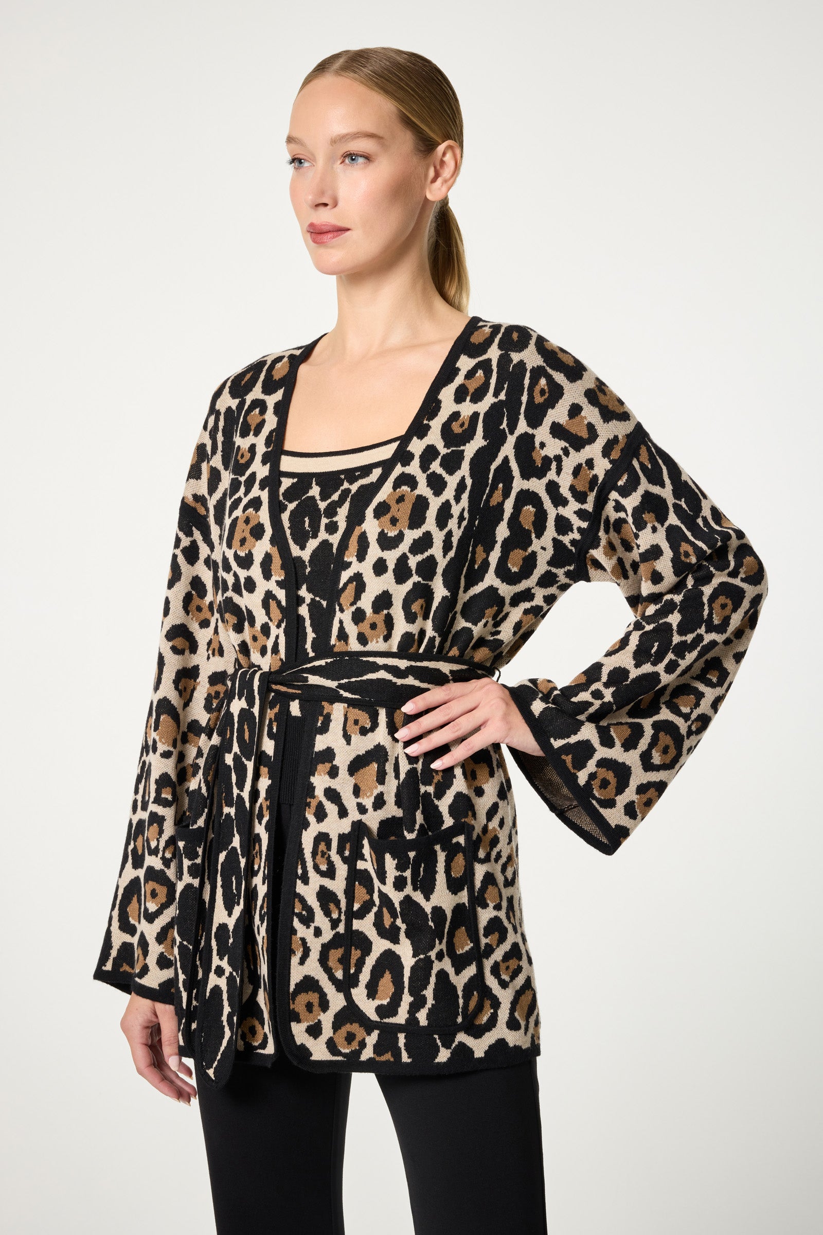 A woman models the Wright Wrap Cardigan, a belted open-front style featuring a bold leopard print, over a matching top. She has straight blonde hair pulled back and pairs the look with black pants against a plain light background.