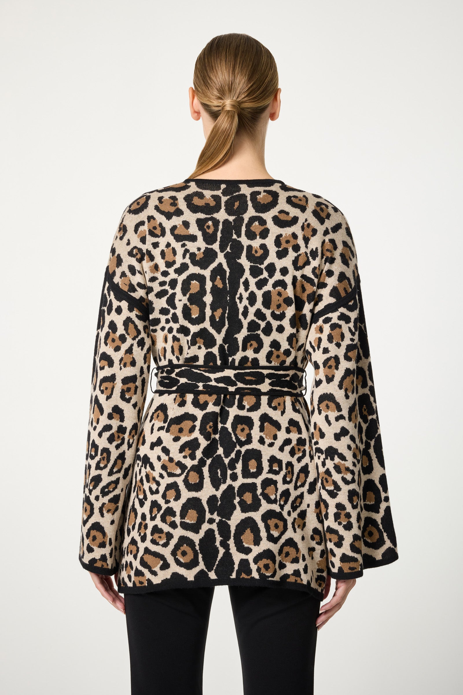 A person with straight, light brown hair in a low ponytail is seen from behind, wearing the Wright Wrap Cardigan—a belted, long-sleeved piece with a bold leopard print—over black pants against a plain white background.