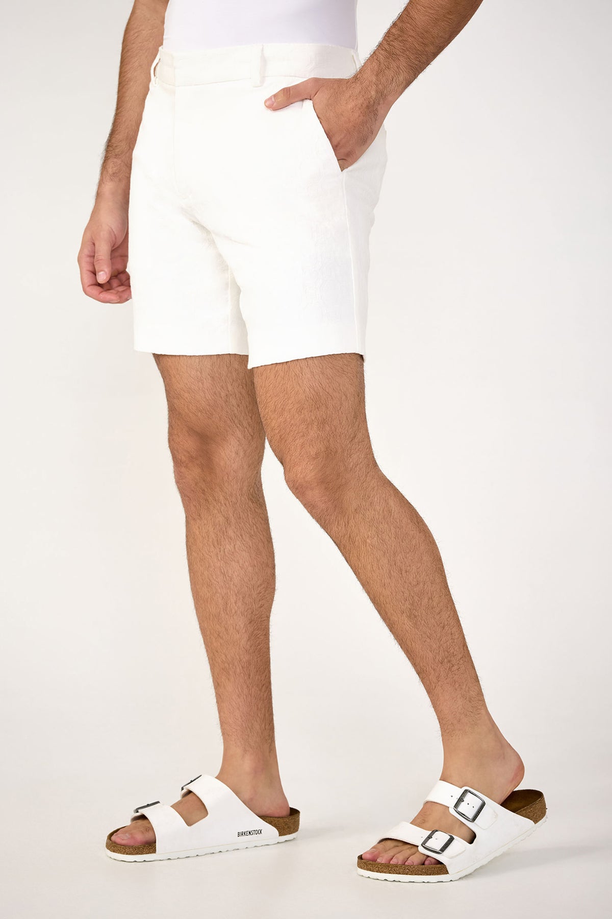 Wearing the Lawrence Short - Stretch Herringbone in white, paired with a matching top and buckled white sandals, a person stands against a plain light background. Only the lower half of their body is visible.