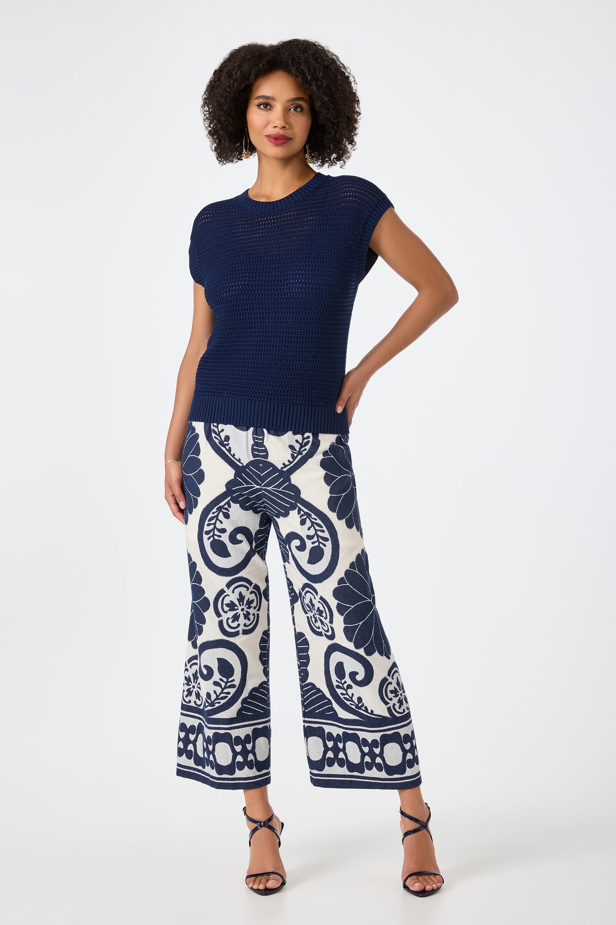 A woman with curly hair confidently poses against a white background wearing the Yuri Knit Pullover, wide-leg white pants with a bold navy floral pattern, and black strappy heels.