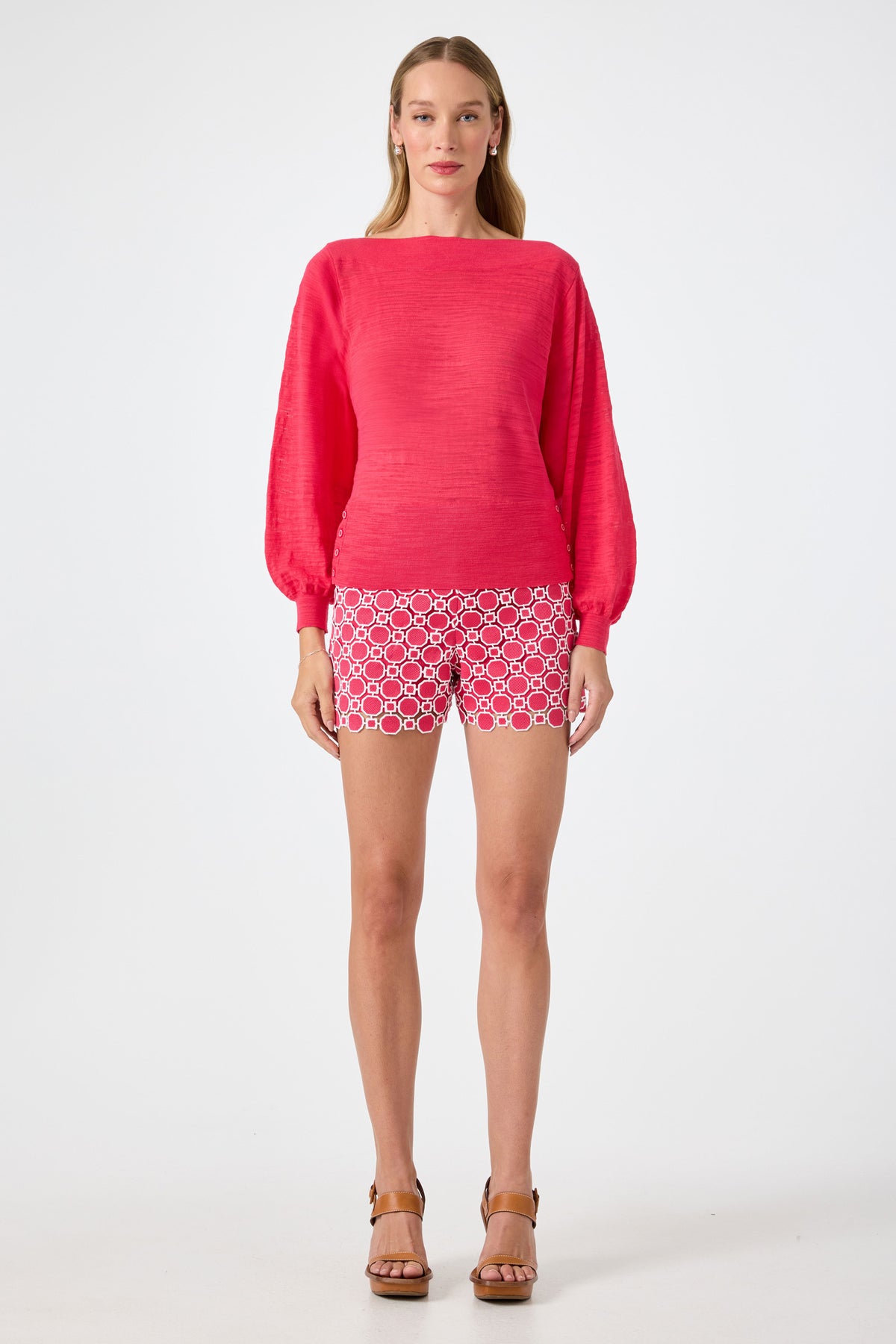 A woman with light brown hair faces forward, wearing the Ramona 2 Boatneck Sweater in bright pink and patterned pink-and-white shorts, paired with tan platform sandals. The background is plain white.