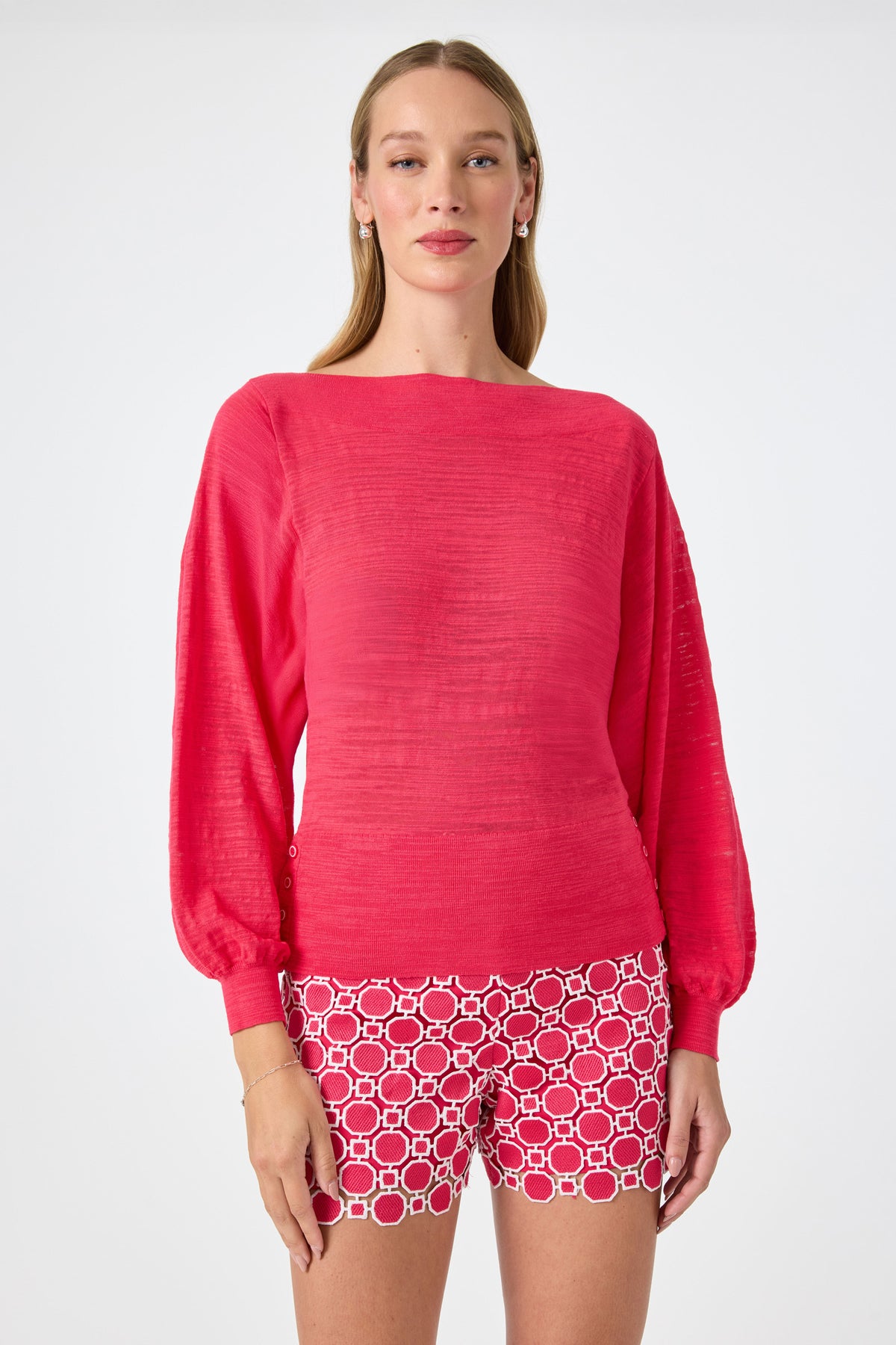 A woman faces forward wearing the Ramona 2 Boatneck Sweater in Cannes Coral. She pairs it with patterned red and white shorts. Her long, straight hair frames a neutral expression against a plain white background.