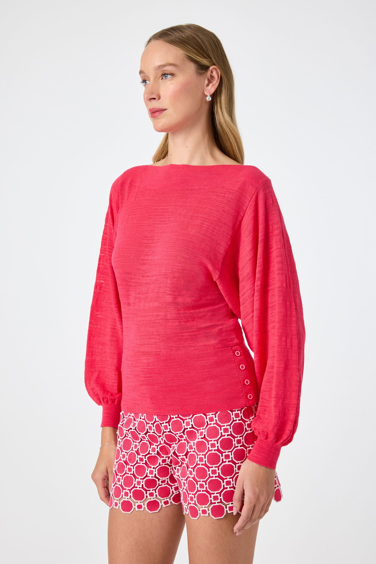 A woman with long blonde hair wears the Ramona 2 Boatneck Sweater, a bright pink long-sleeve top with balloon sleeves, paired with patterned red-and-white shorts, standing against a plain white background.
