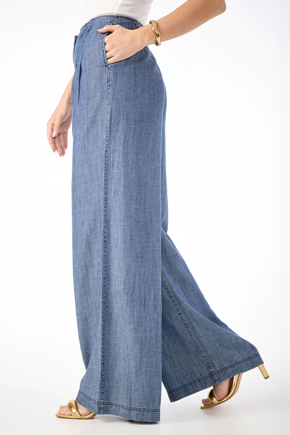 Wearing Dans Wide-Leg Chambray Pants, a white top, a gold bracelet, and gold open-toe heels, a person stands against a white background with one hand in their pocket and the other by their side.