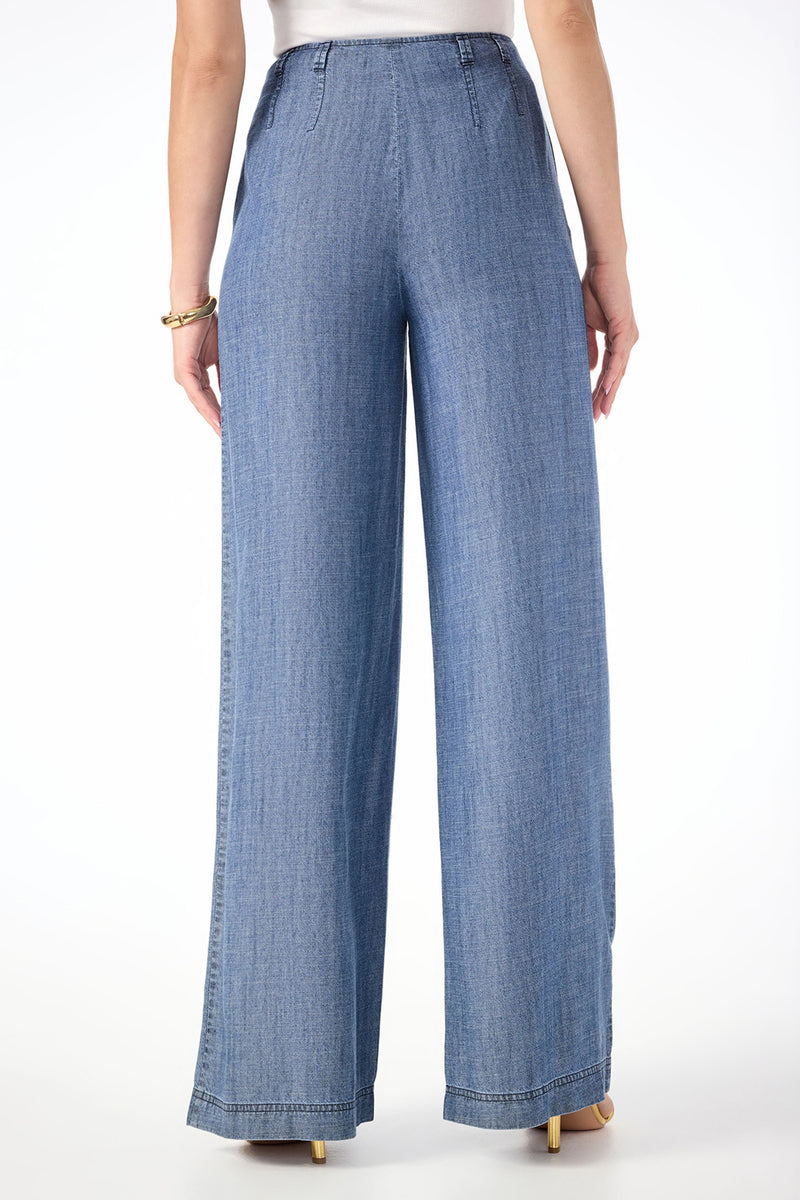 A person is shown from the back wearing Dans Wide-Leg Chambray Pants, which are high-waisted, loose-fitting, and long. The blue denim pants are styled with a white top and beige heeled shoes.