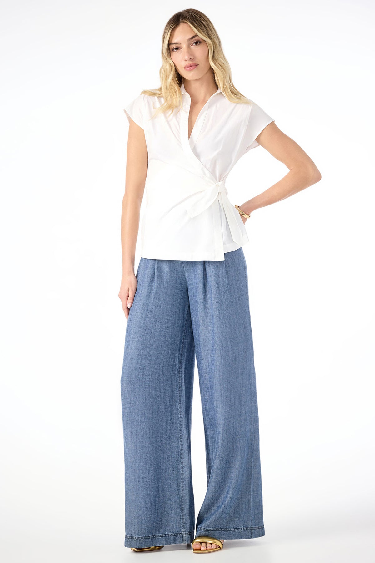 A woman stands against a white background wearing a short-sleeved wrap blouse with a side tie and Dans Wide-Leg Chambray Pants. She has long, wavy blonde hair and is wearing sandals.