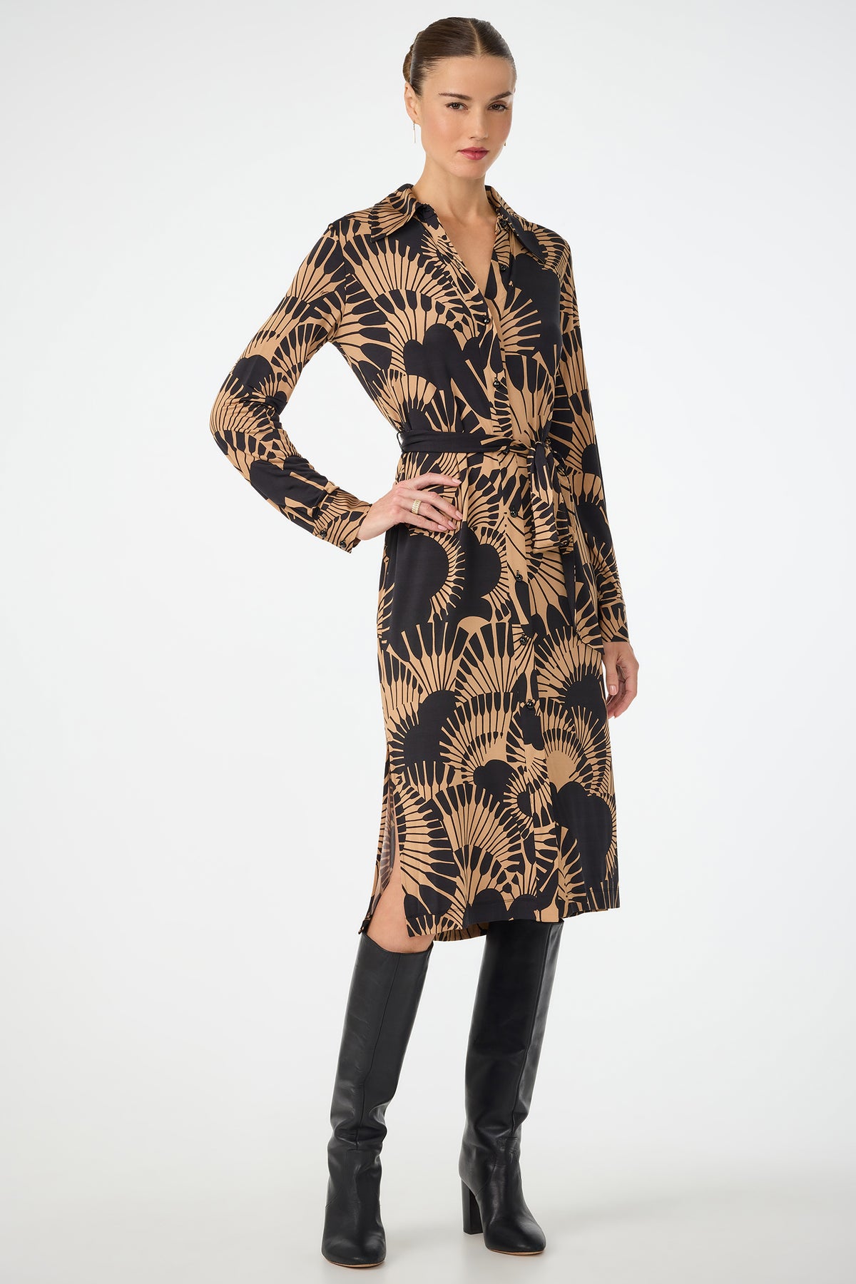 A woman stands confidently in the Radio City Wrap Dress - Juniper Jersey, a knee-length black dress with tan botanical prints, collared neckline, and waist tie. She pairs it with tall black heeled boots against a plain white background.
