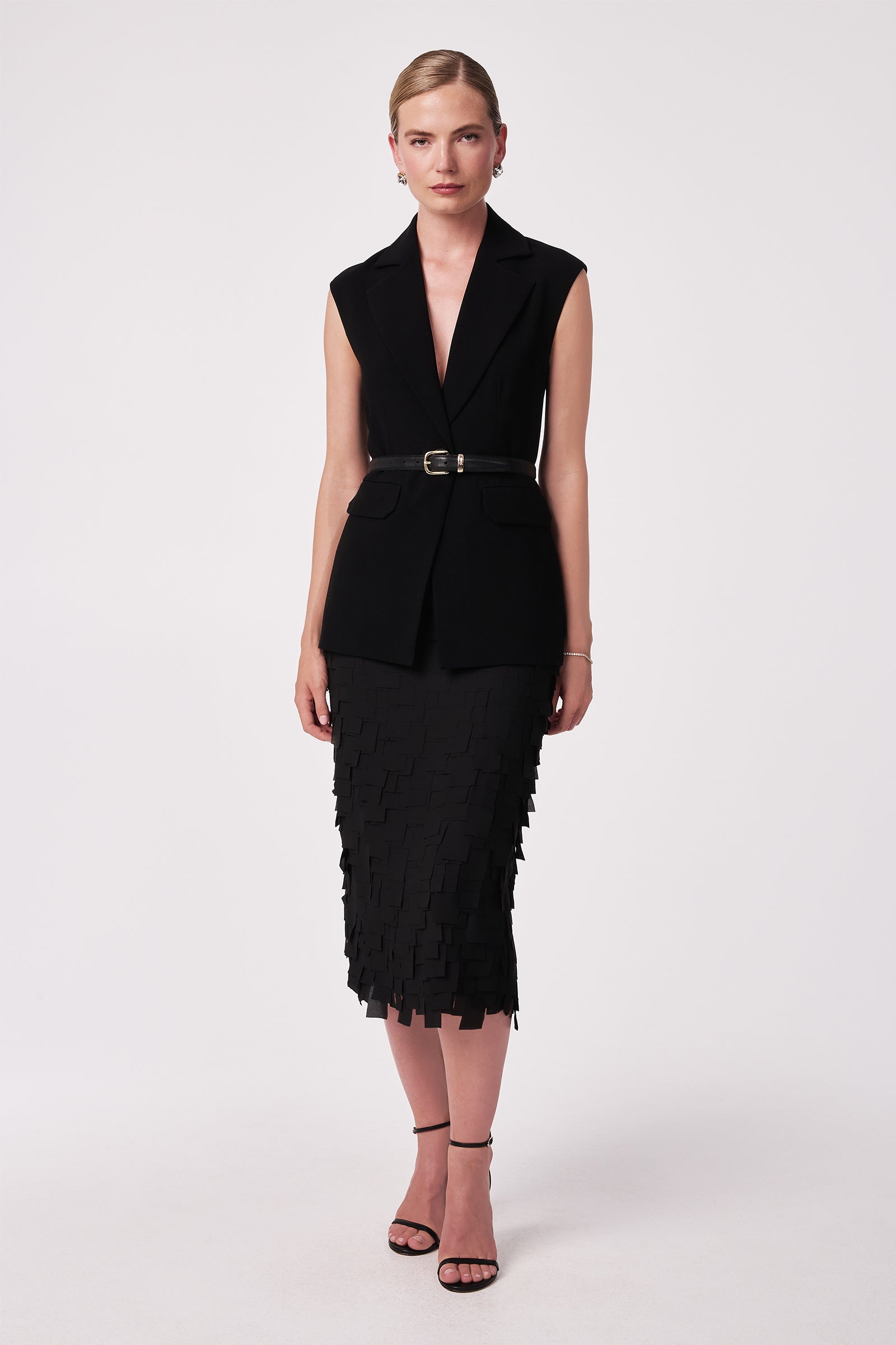 A woman stands against a plain background, wearing a sleeveless black belted blazer and the Dita Midi Pencil Skirt - Flutter Chiffon. Her hair is pulled back and she completes the look with black strappy heels.
