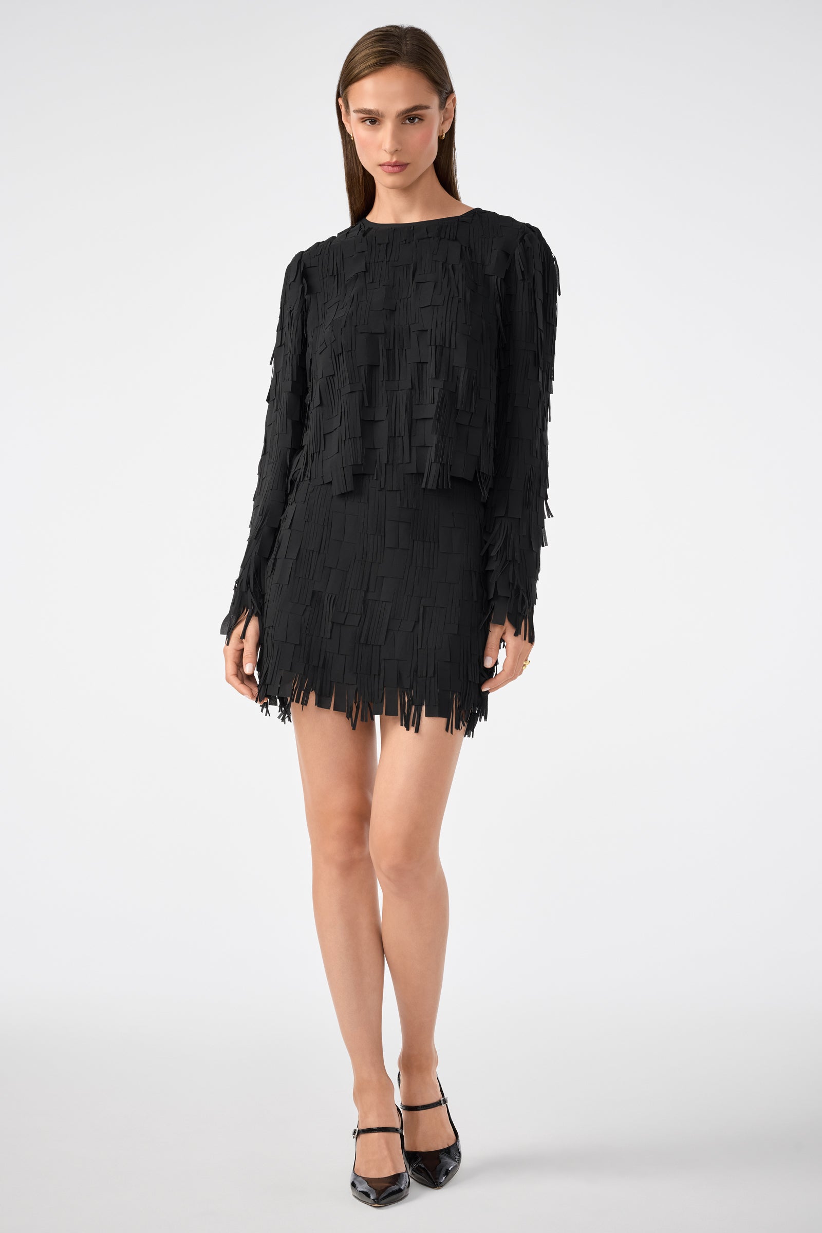 A woman stands against a white background wearing the Chinaberry Top, a black, long-sleeve, textured mini dress with rectangular fringe details, paired with black pointed-toe heels.