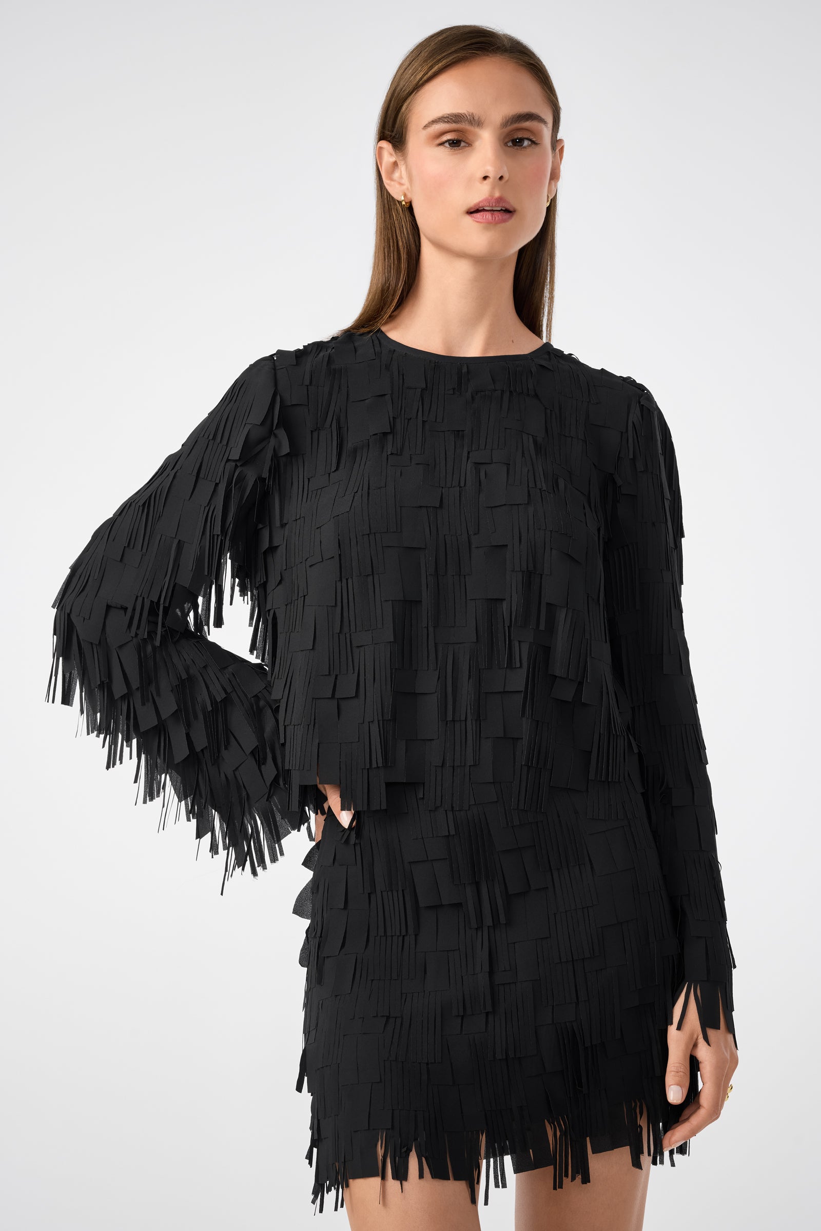 A woman with straight brown hair wears the Chinaberry Top—a black, long-sleeve piece featuring layered rectangular fringe—while standing against a plain light background.