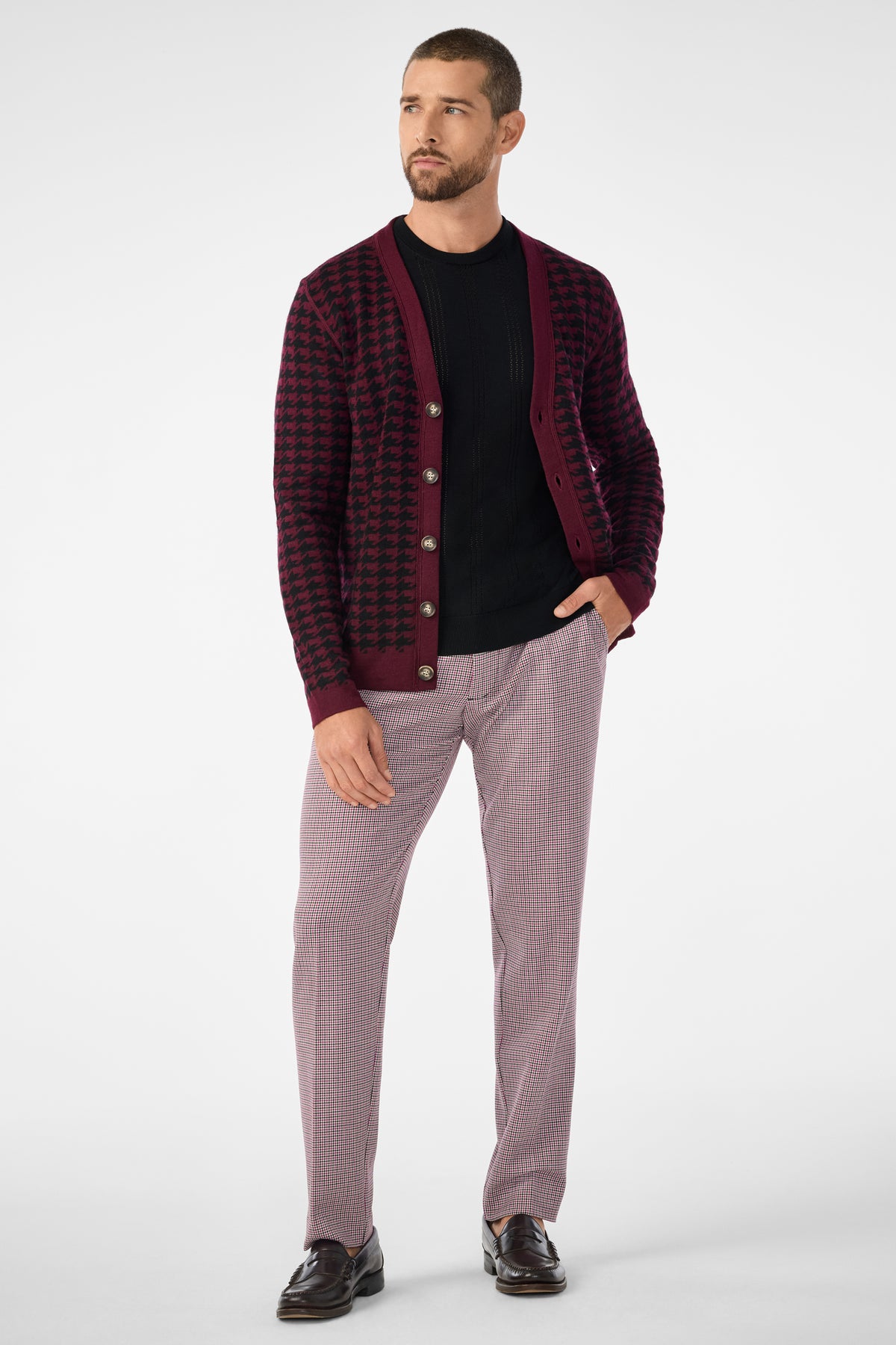 A man with short hair and a beard stands against a plain background, wearing the Alder Cardigan in a burgundy and black pattern over a black shirt, light-patterned trousers, and dark loafers, looking to his left.