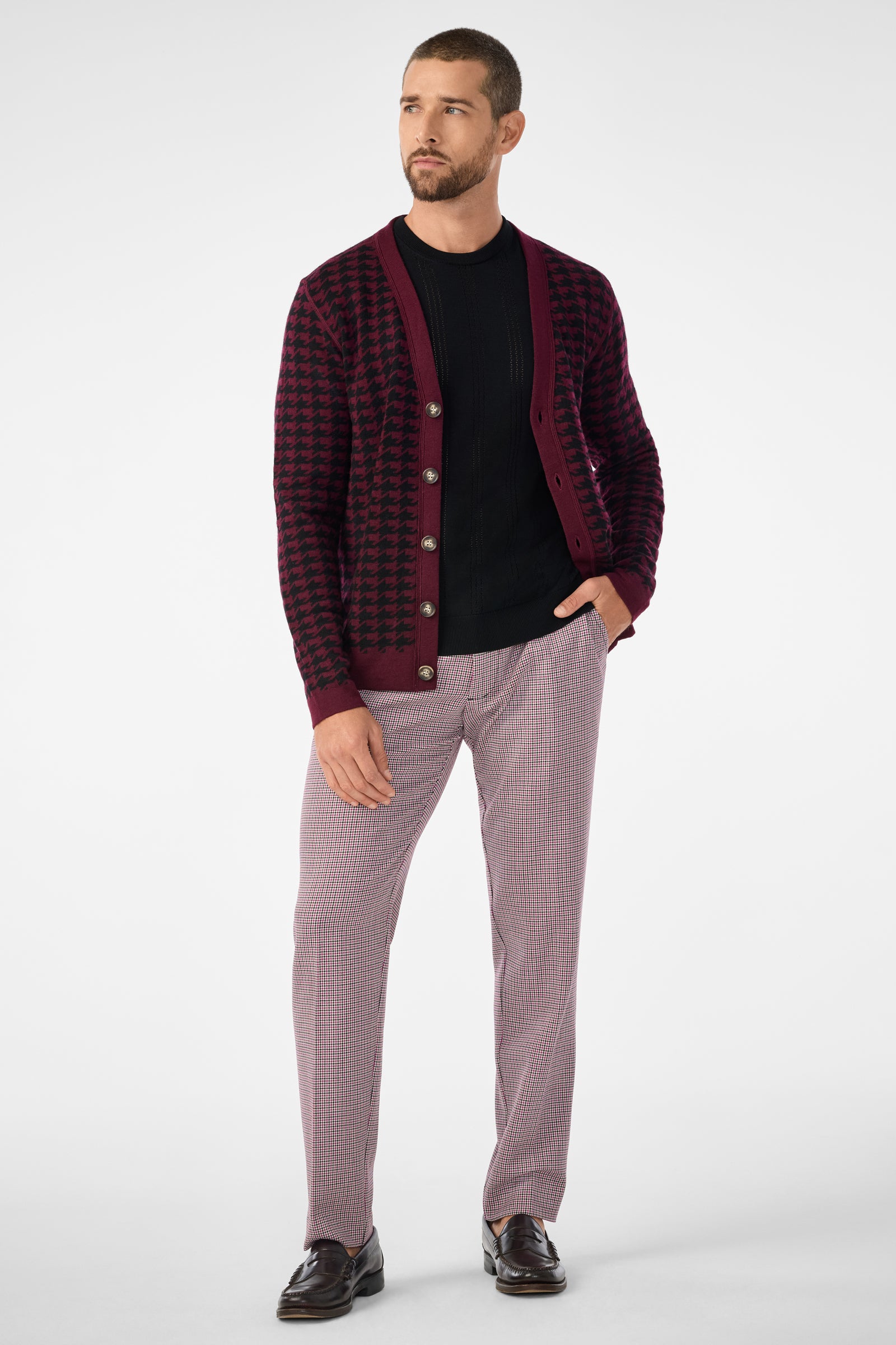 A man with short hair and a beard stands against a plain background, wearing the Alder Cardigan in a burgundy and black pattern over a black shirt, light-patterned trousers, and dark loafers, looking to his left.