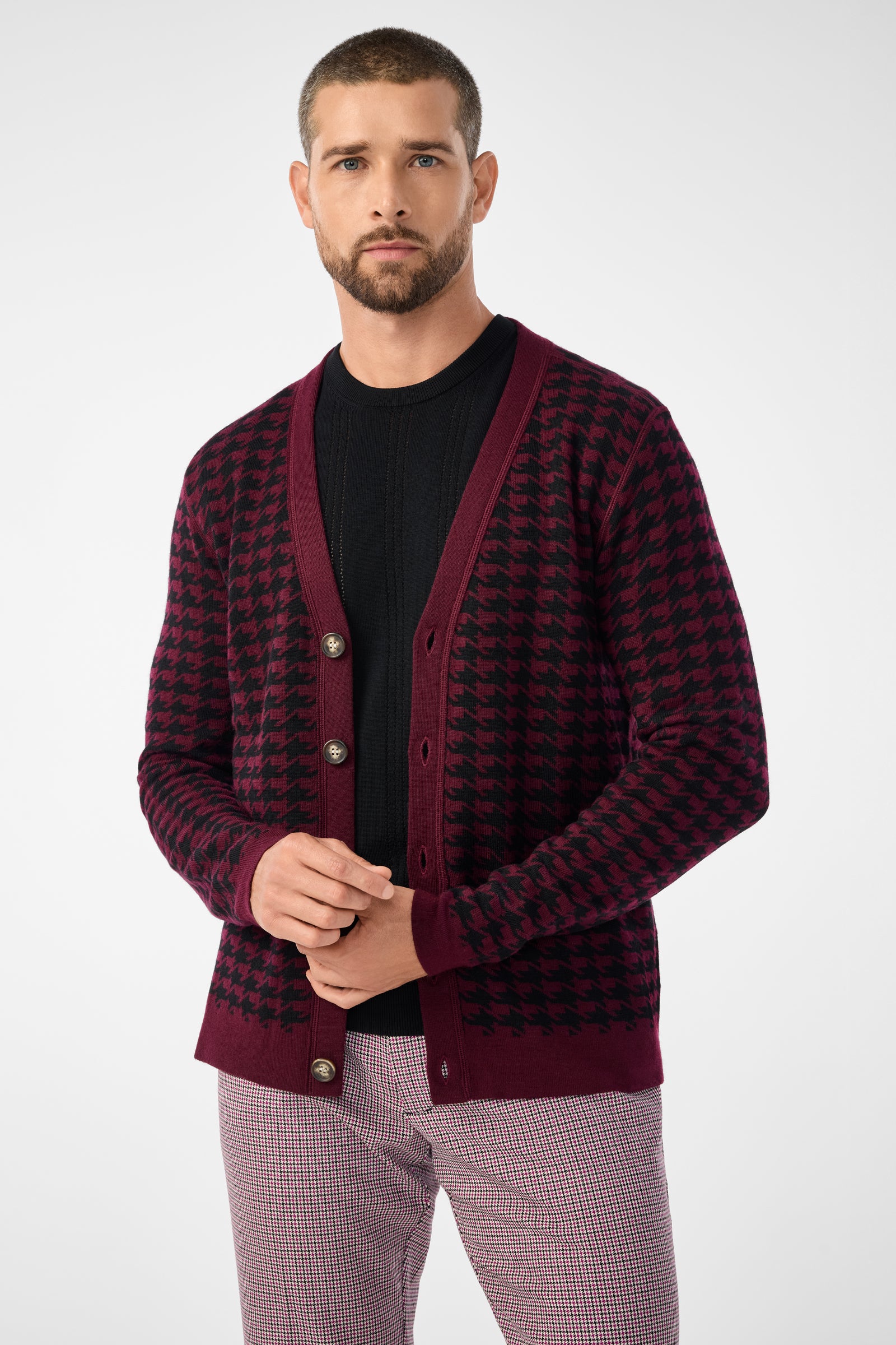 A man with short hair and a beard wears the Alder Cardigan—a maroon and black houndstooth sweater—over a black shirt and light plaid pants, standing against a plain light background.