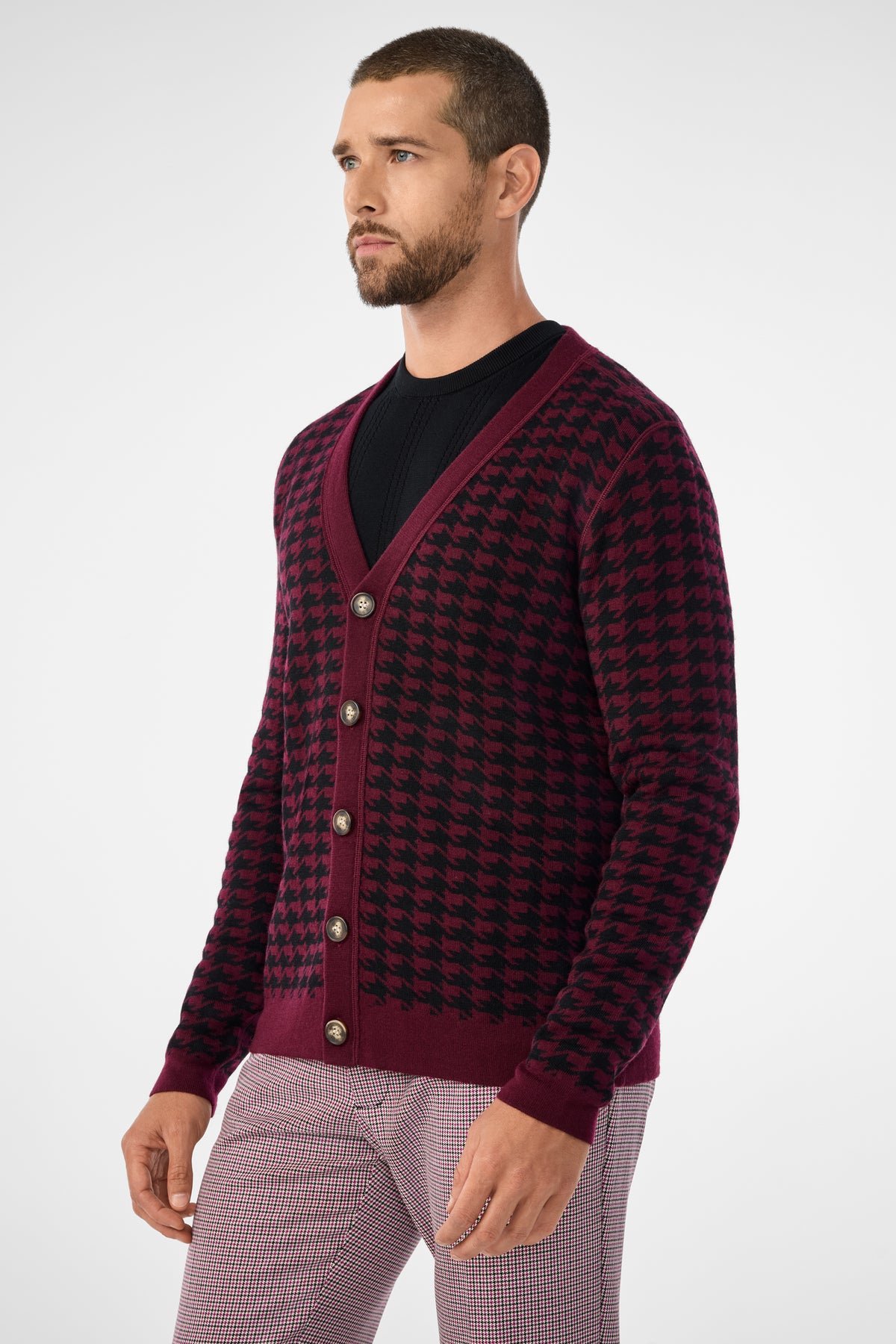 A man with a beard wears the Alder Cardigan, a burgundy and black houndstooth style with large buttons, over a black shirt and light plaid pants. He stands facing slightly left against a plain light background.