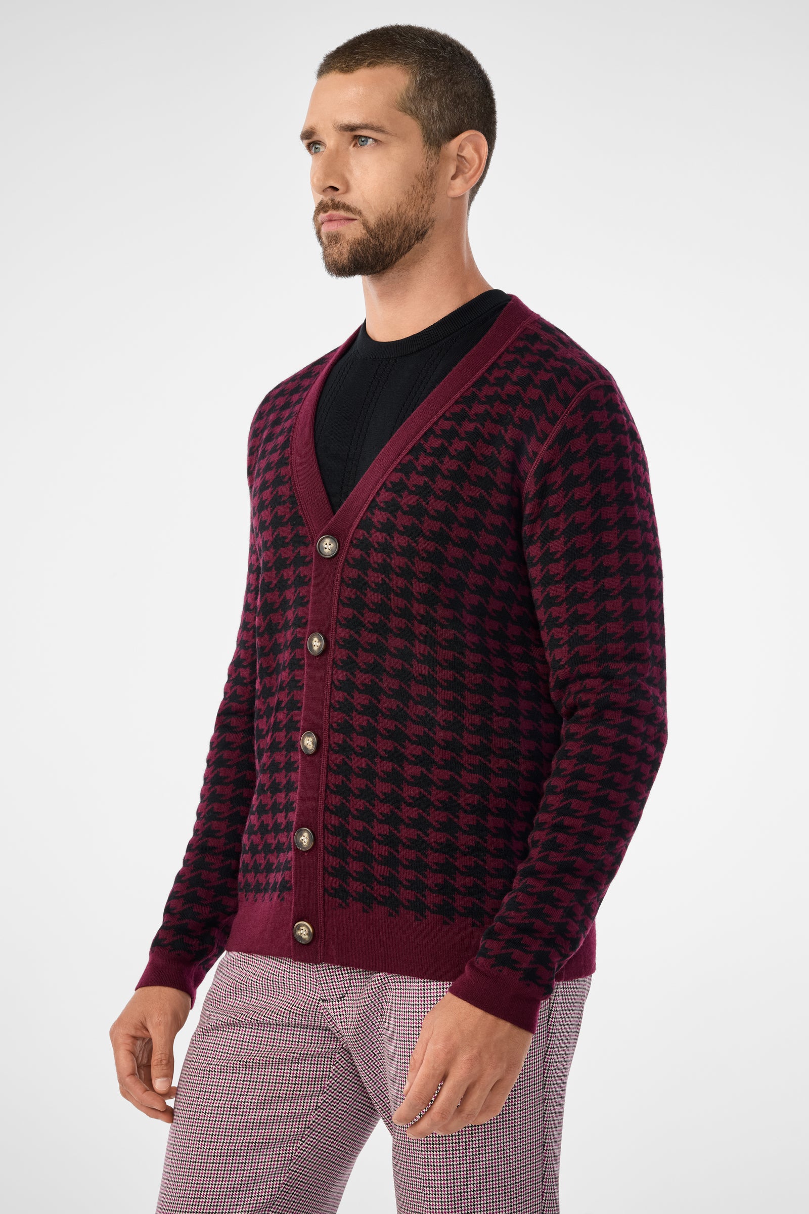 A man with a beard wears the Alder Cardigan, a burgundy and black houndstooth style with large buttons, over a black shirt and light plaid pants. He stands facing slightly left against a plain light background.