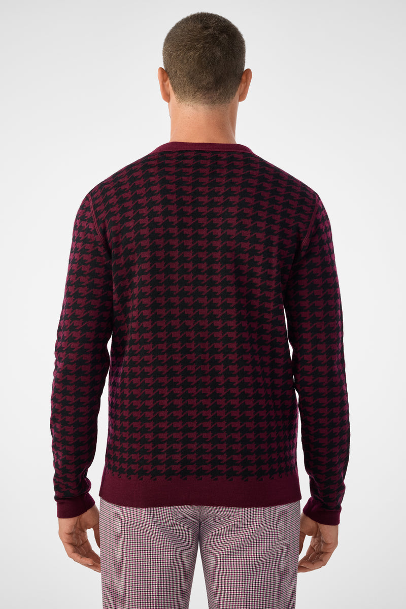 A man is seen from behind wearing the Alder Cardigan, featuring a burgundy and black houndstooth pattern, paired with light plaid pants against a plain light background.