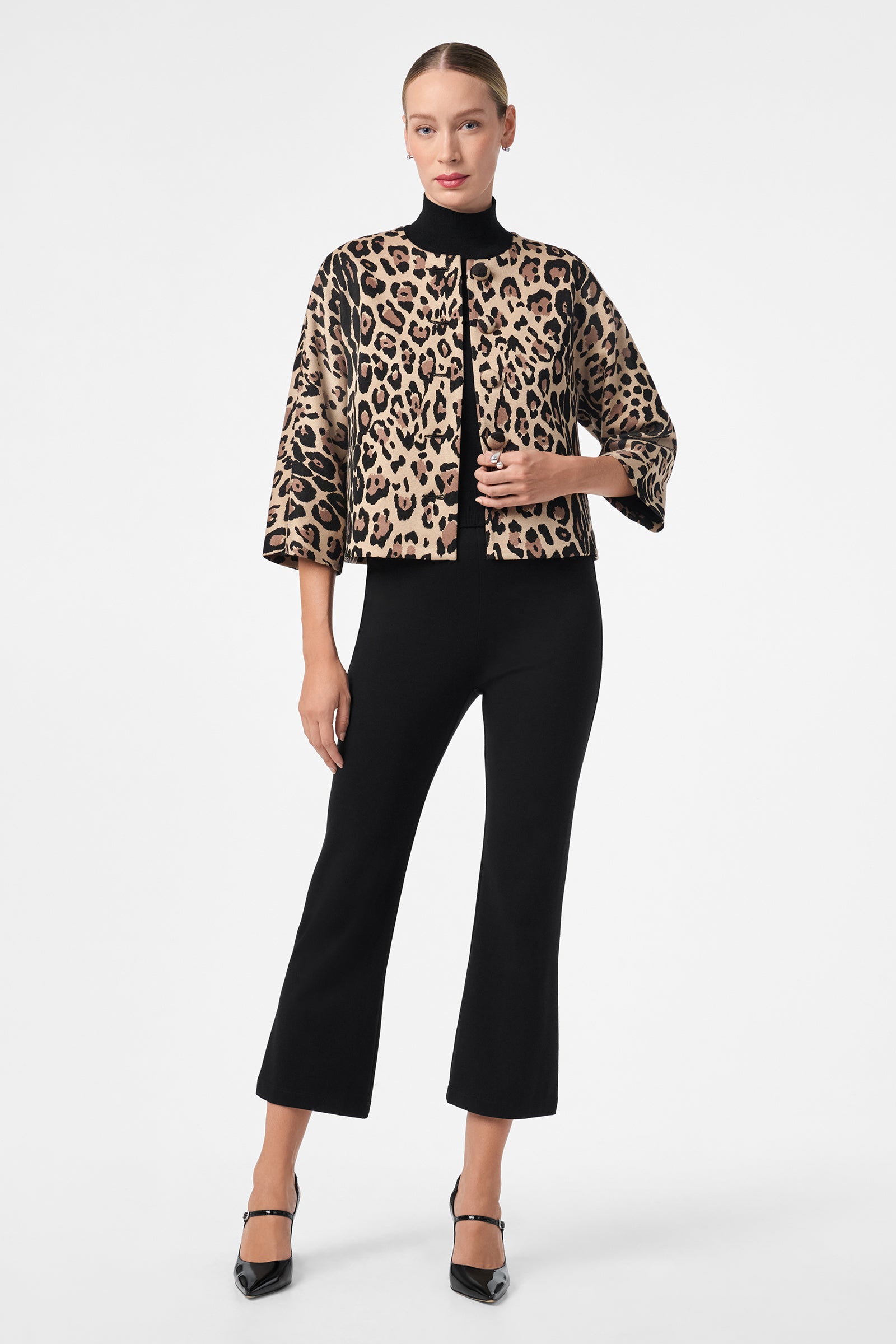A woman with her hair pulled back and a neutral expression stands against a plain white background wearing the Critique Jacket—an animal print, cropped style with black trim—paired with black wide-leg pants and heels.