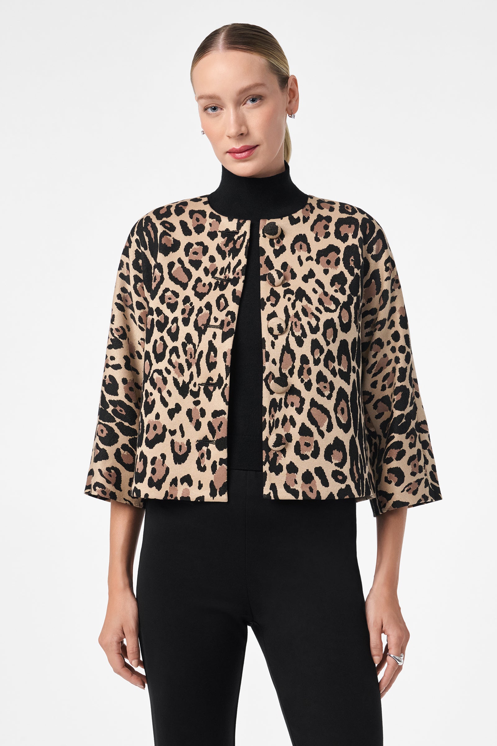 A woman stands against a plain white background, wearing black pants and a black turtleneck topped with the Critique Jacket—a cropped, open-front jacket with a bold leopard print pattern.