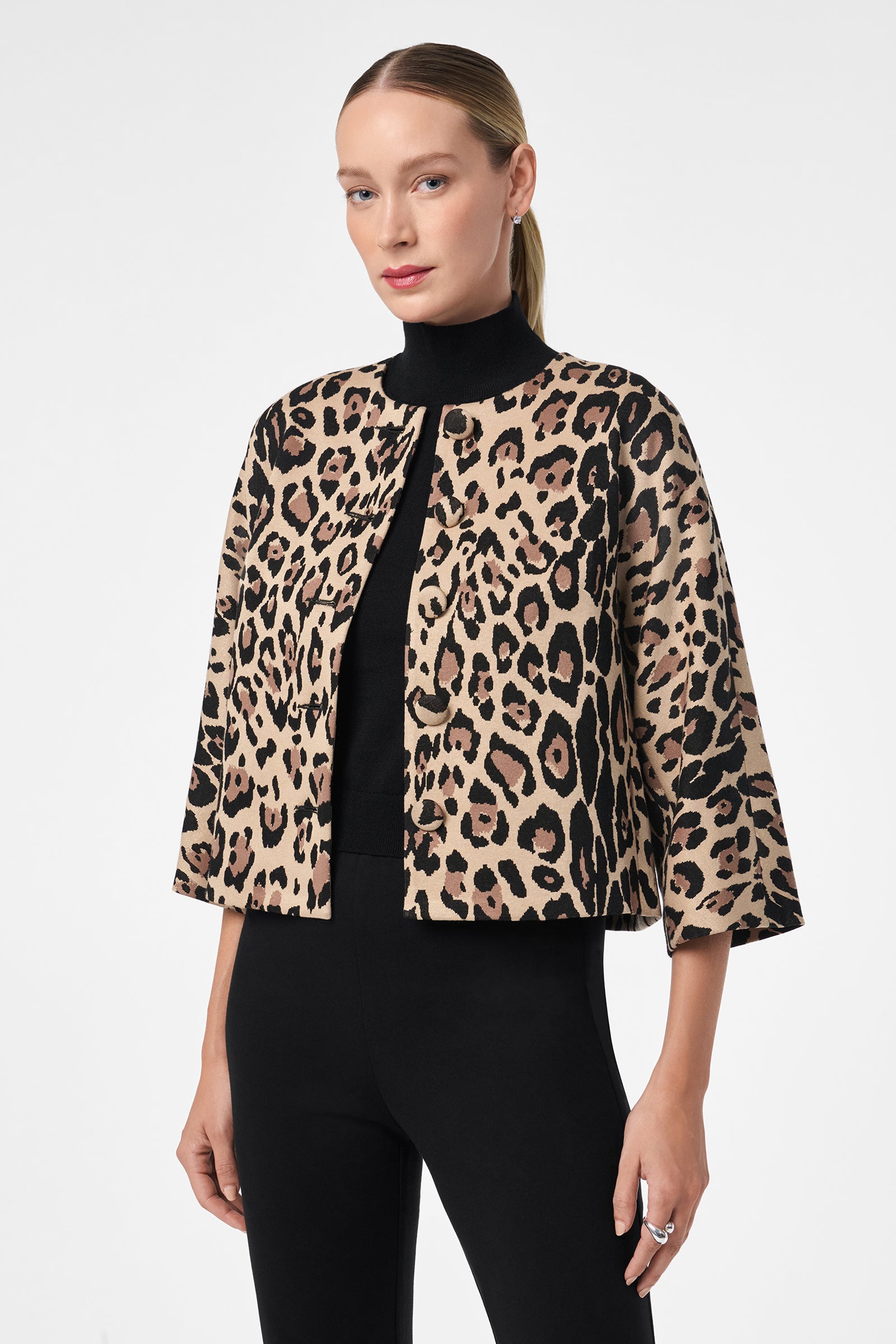 A woman with straight, pulled-back blonde hair wears a black turtleneck and pants, topped with the Critique Jacket—a cropped, open-front piece in bold leopard print. She stands against a plain white background.