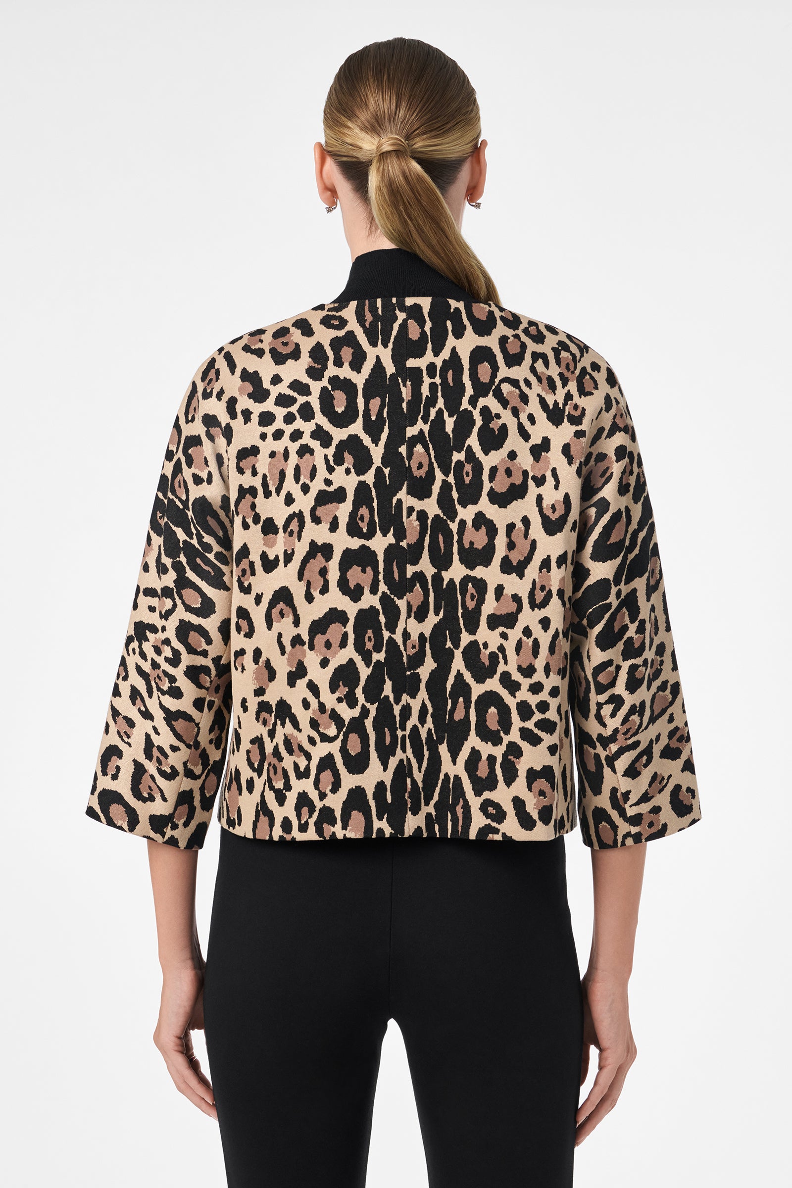A woman with straight, light brown hair in a low ponytail models the Critique Jacket—a cropped, leopard-print piece with three-quarter sleeves—paired with black fitted pants, photographed from the back against a plain white background.