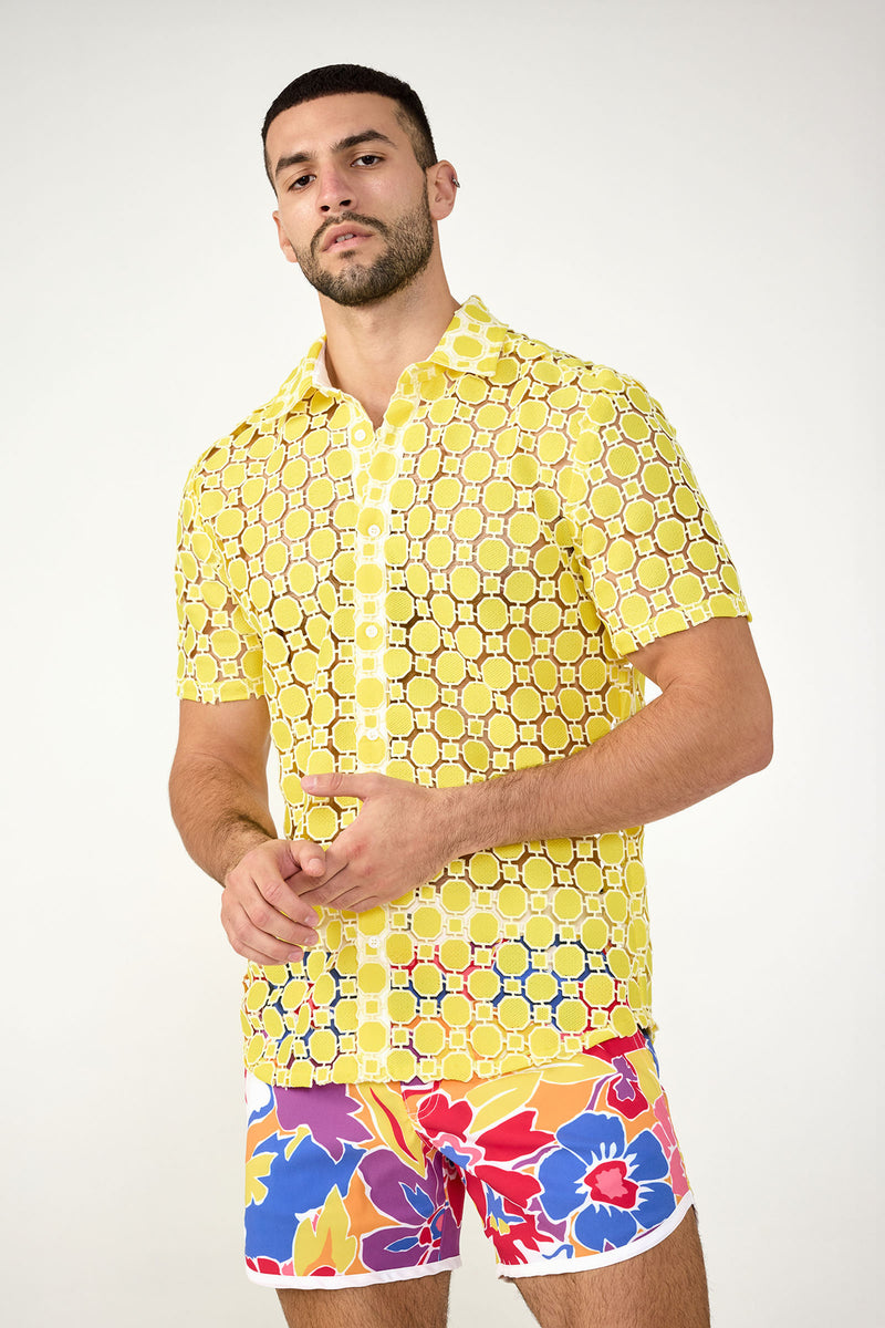 A man stands confidently against a plain background, wearing the Long Beach Short-Sleeve Shirt in Lavandou Lemon with a sheer geometric pattern, paired with colorful floral shorts.