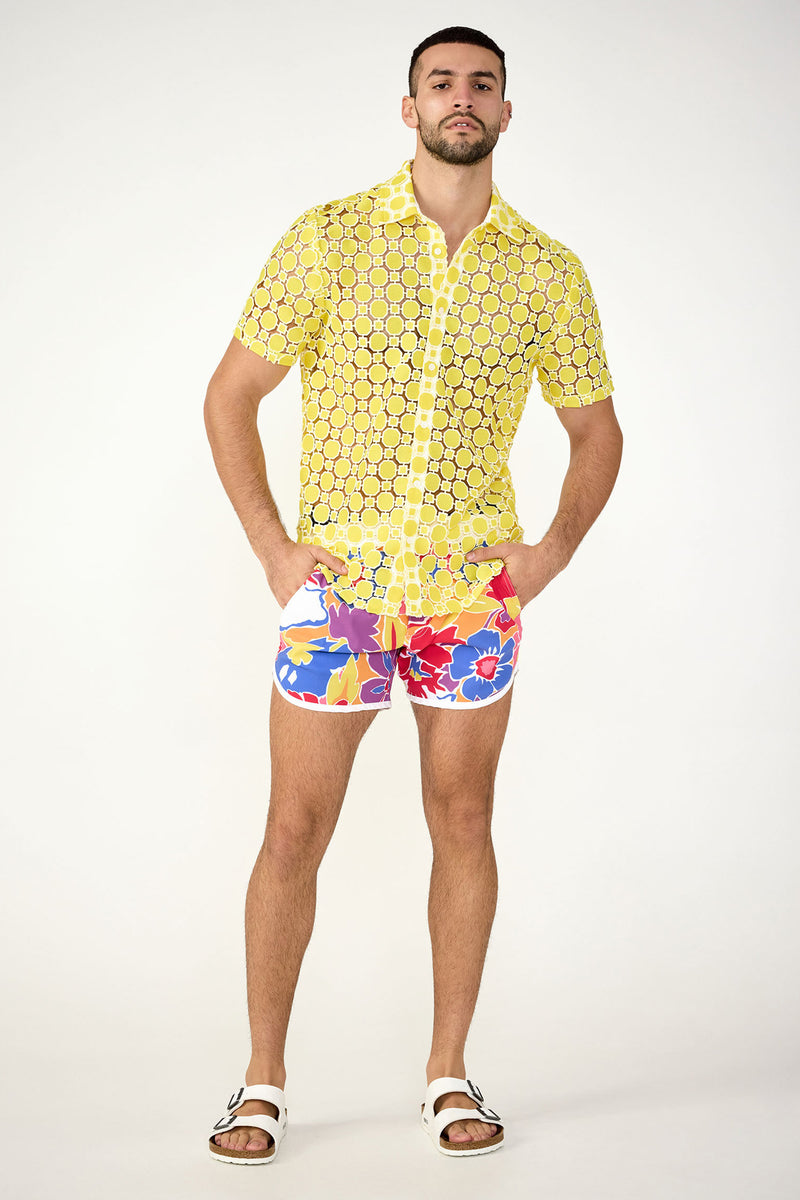 A man with a neutral expression stands against a plain background wearing the Long Beach Short-Sleeve Shirt, colorful floral print shorts, and white sandals. His hands are in his pockets.