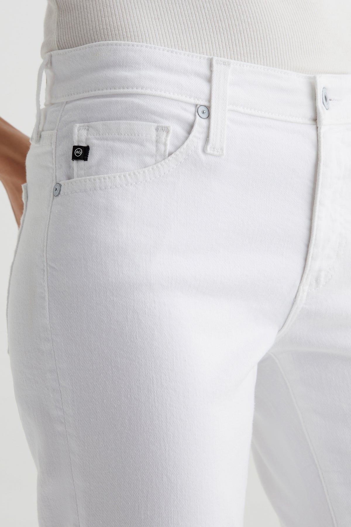 Close-up of a person wearing AG Angel Low-Rise Bootcut Jean in white and a matching top, featuring a small black logo tag on the front pocket. Focus is on the waist and upper thigh area.