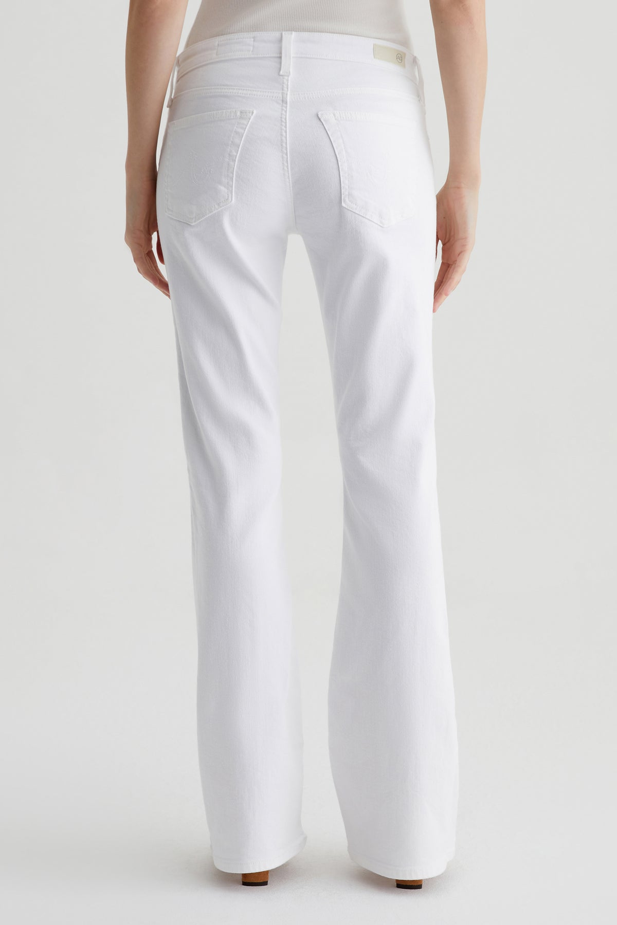 A person stands facing away from the camera, wearing the AG Angel Low-Rise Bootcut Jean in white and a light-colored top, set against a plain white background.