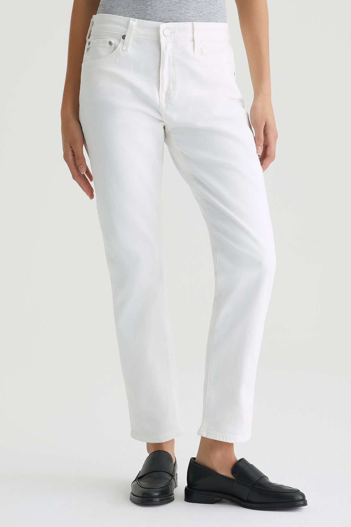 A person wears AG Mid-Rise Slouchy Ex Boyfriend Jeans in white with a light gray top and black loafers, standing against a plain light background.