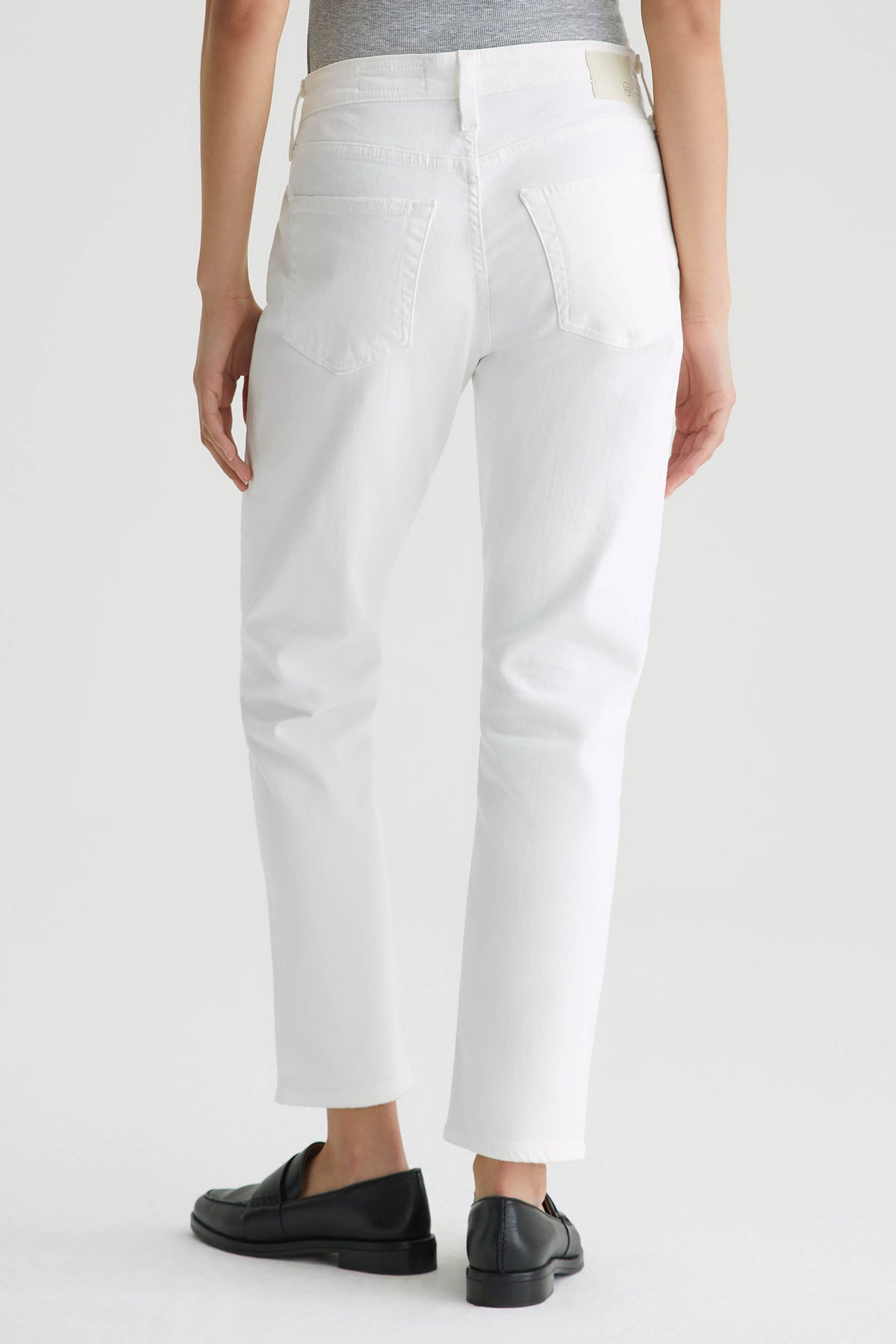 Person stands facing away in a gray top tucked into AG Mid-Rise Slouchy Ex Boyfriend Jean - White with black loafers; the image highlights the relaxed fit and back view of the jeans against a plain background.