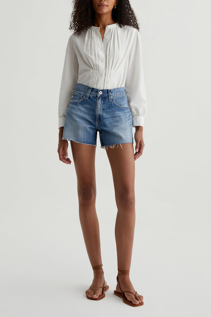 A person wearing a white long-sleeve blouse, AG Halle Denim Short - Blue, and brown flat sandals stands against a plain light background.