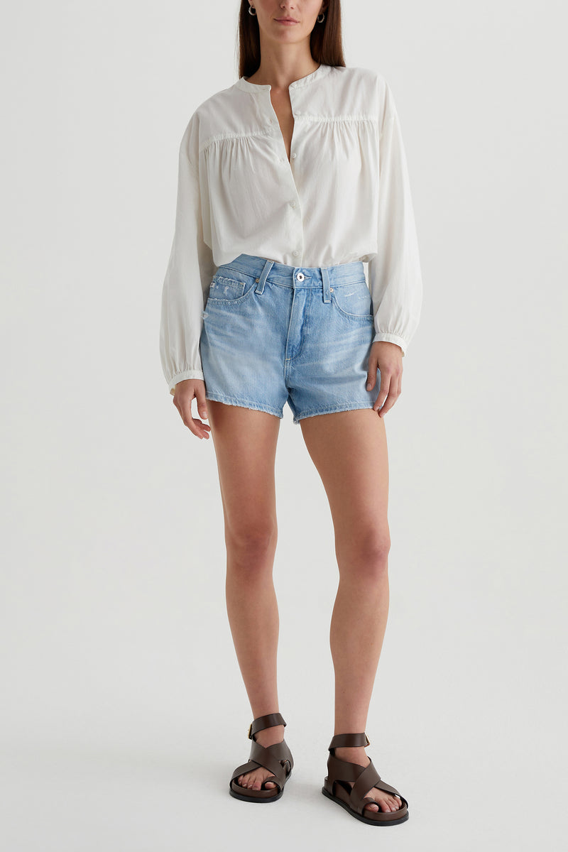 A woman stands facing forward against a plain white background, wearing a loose white long-sleeve blouse, dark brown sandals, and the AG Halle Denim Short in Light Blue. Her hair is down.