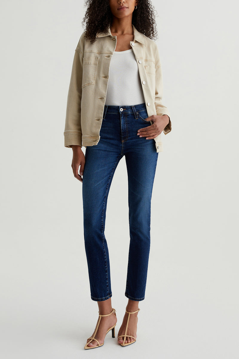 A woman stands against a plain background wearing the AG Mari Slim-Straight Crop Jean in indigo, a beige denim jacket, white top, and beige heeled sandals. She lightly holds the open jacket, with her head not fully visible.