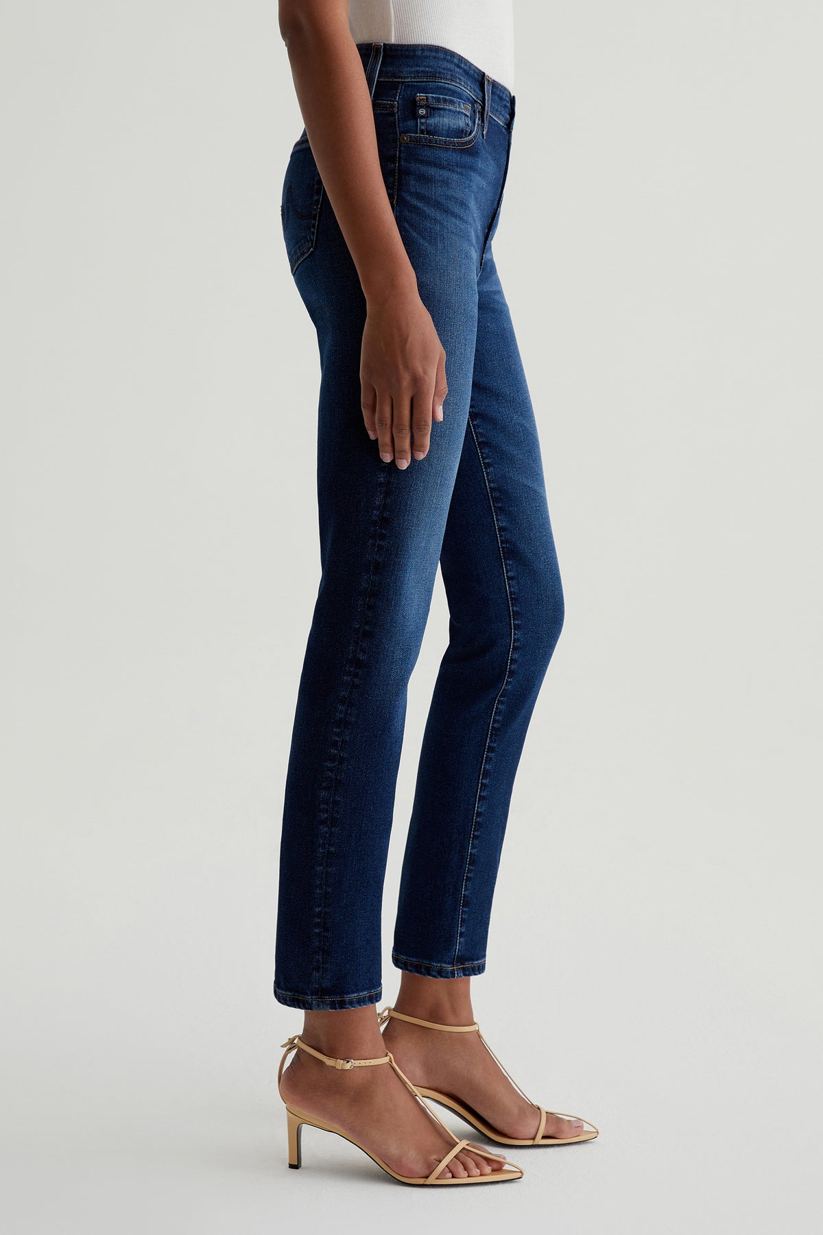 A person models the AG Mari Slim-Straight Crop Jean in Indigo with a white top and beige heeled sandals, shown from shoulder to feet in a side view against a plain light background.