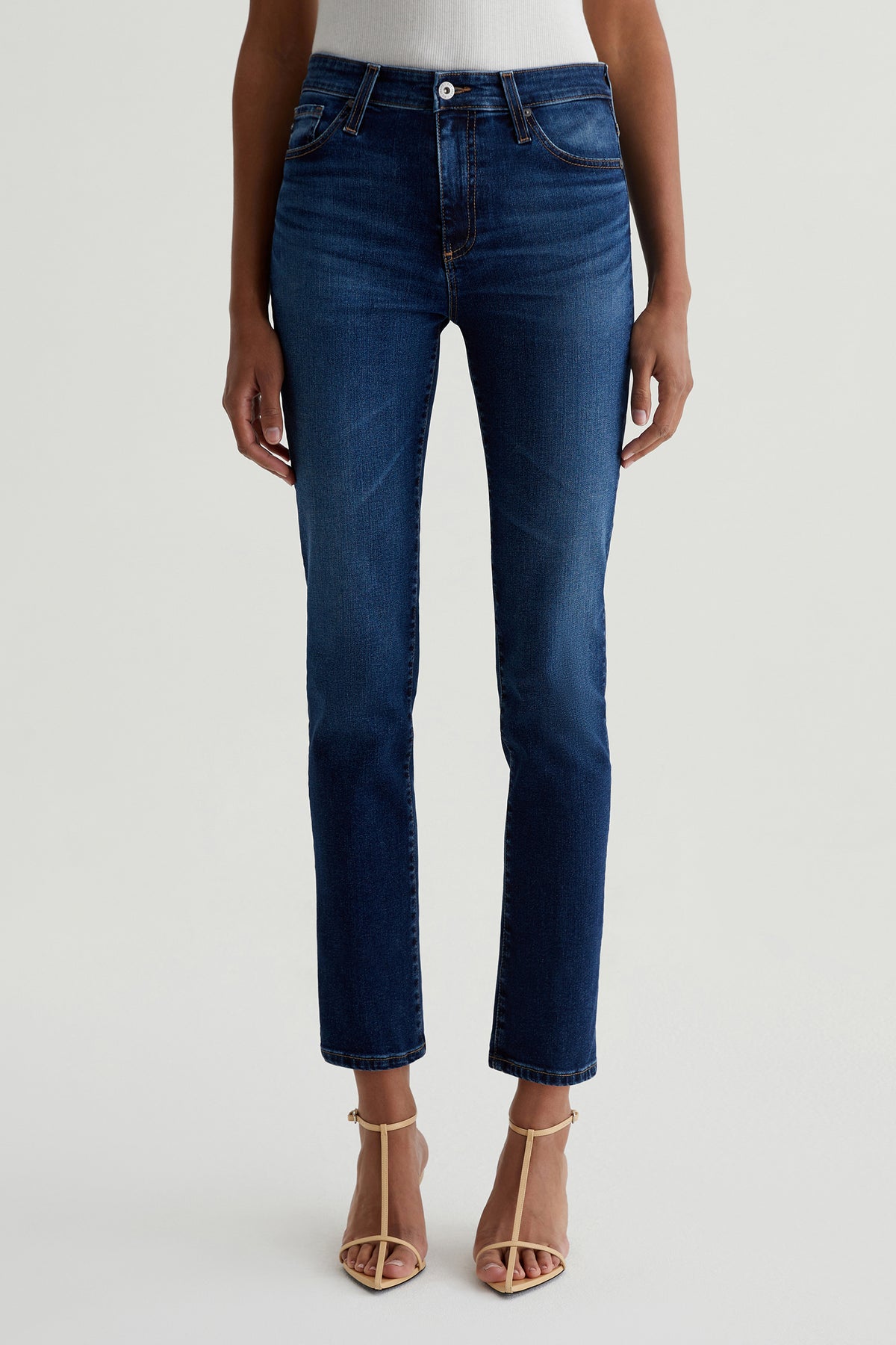 A person stands against a plain light background wearing AG Mari Slim-Straight Crop Jean in indigo with beige strappy heels and a white top tucked in; the upper body is partially visible.