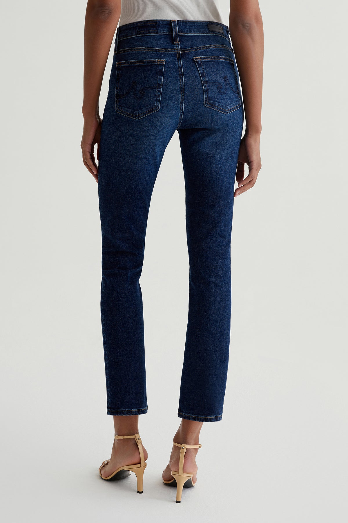 Someone is shown from the back wearing AG Mari Slim-Straight Crop Jeans in indigo and beige high-heeled sandals, standing against a plain light background.