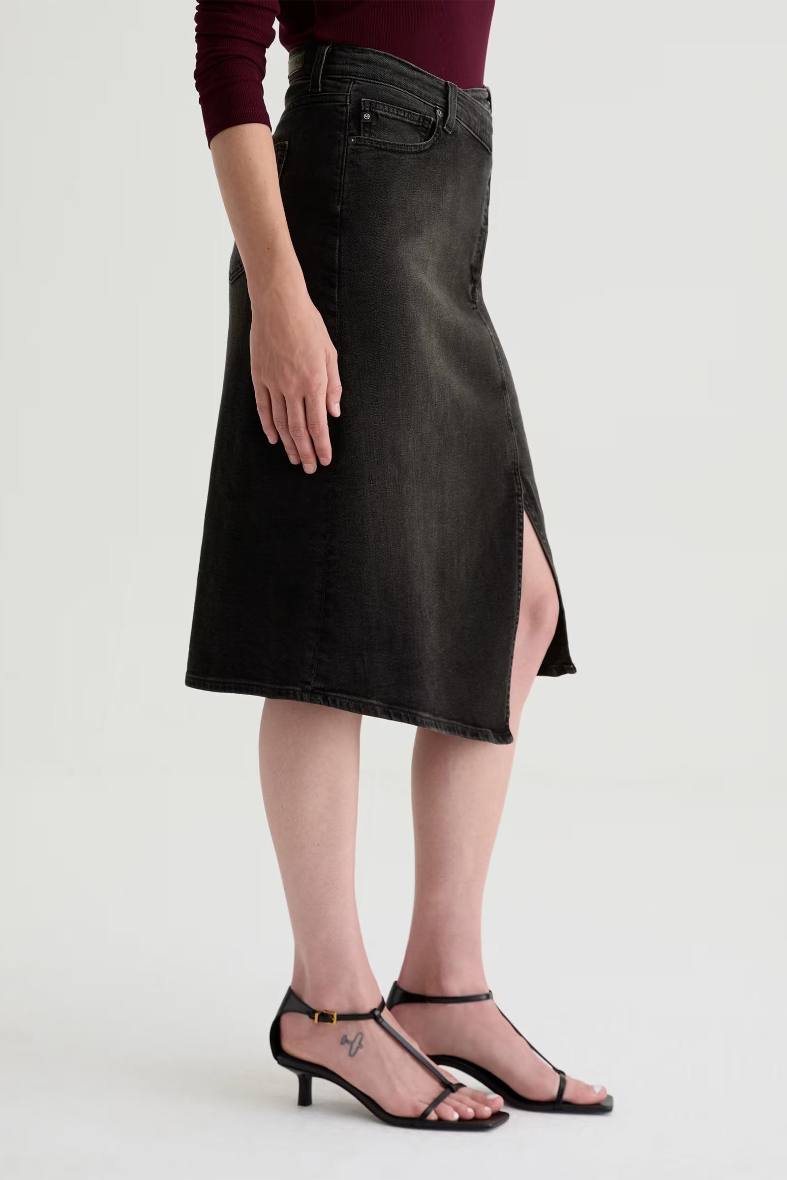 A person wearing the Bruna Wrap Midi Skirt in dark denim with a front slit, paired with a maroon long-sleeve top and black strappy heeled sandals, stands against a plain light background.